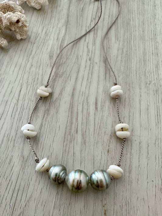 a necklace with three silvery tahitain pearls on a gry silk thread with white small puka shells  is on a light grey wooden background. there is a small chink of coral in the upper left corner