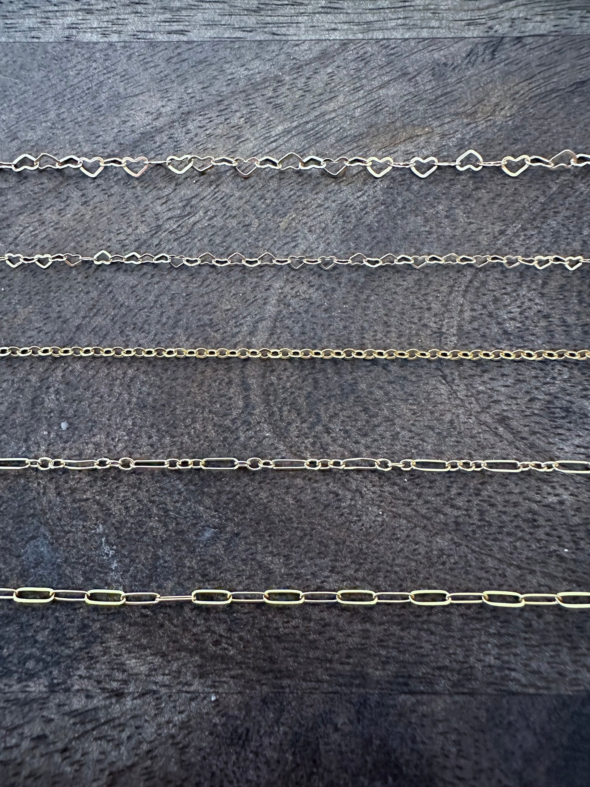 five different chains laid out in a row on a grey back ground. the firs chain is made by linked hearts, the second is smaller linked hearts, third is a small circular chain, the fourth has one long link then three circular links in an alternating pattern, the fifth is a paperclip chain all long links