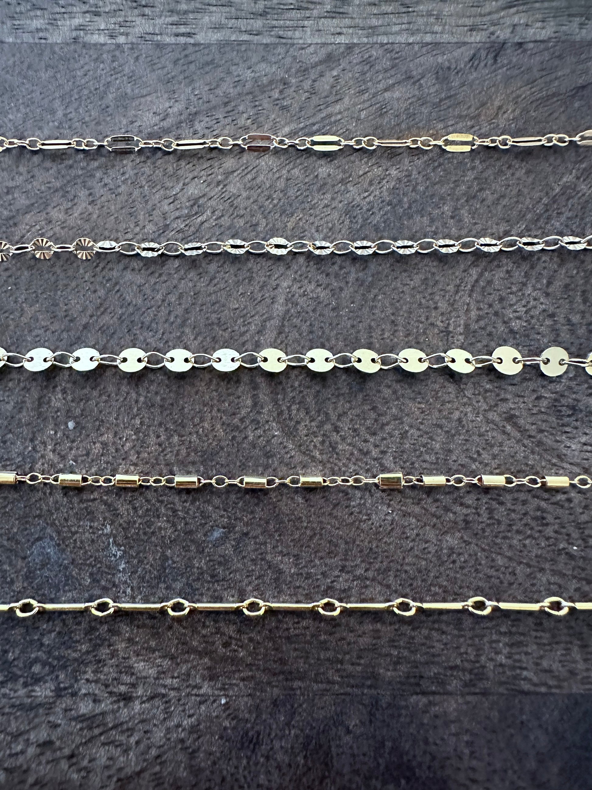 five chaings on a wooden background. the first has flat oval link with two round links connecting it. the oval has a slit int he center. the second chain has alternating round discs with an embosssed pattern and oval links. the third chain is flat round discs alternating wiht a twisted oval link. the fourth is a tube of gold linked by three small round links. the fifth is one long bar linked with small ovals.