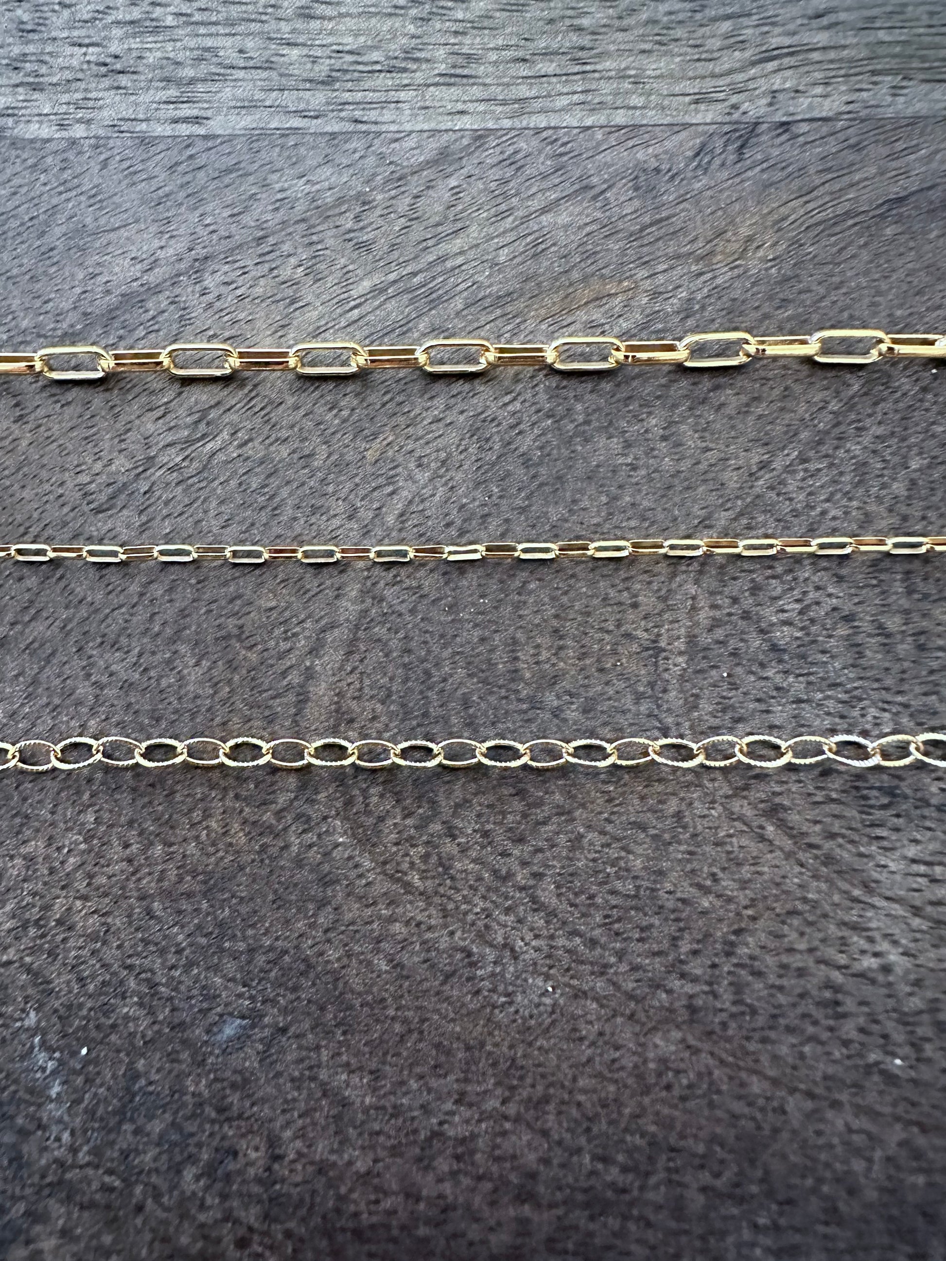 three chains on a grey wooden background. the first chain is large rectangular links, the second is smaller rectangular links, the third is alternating textured and polished oval links