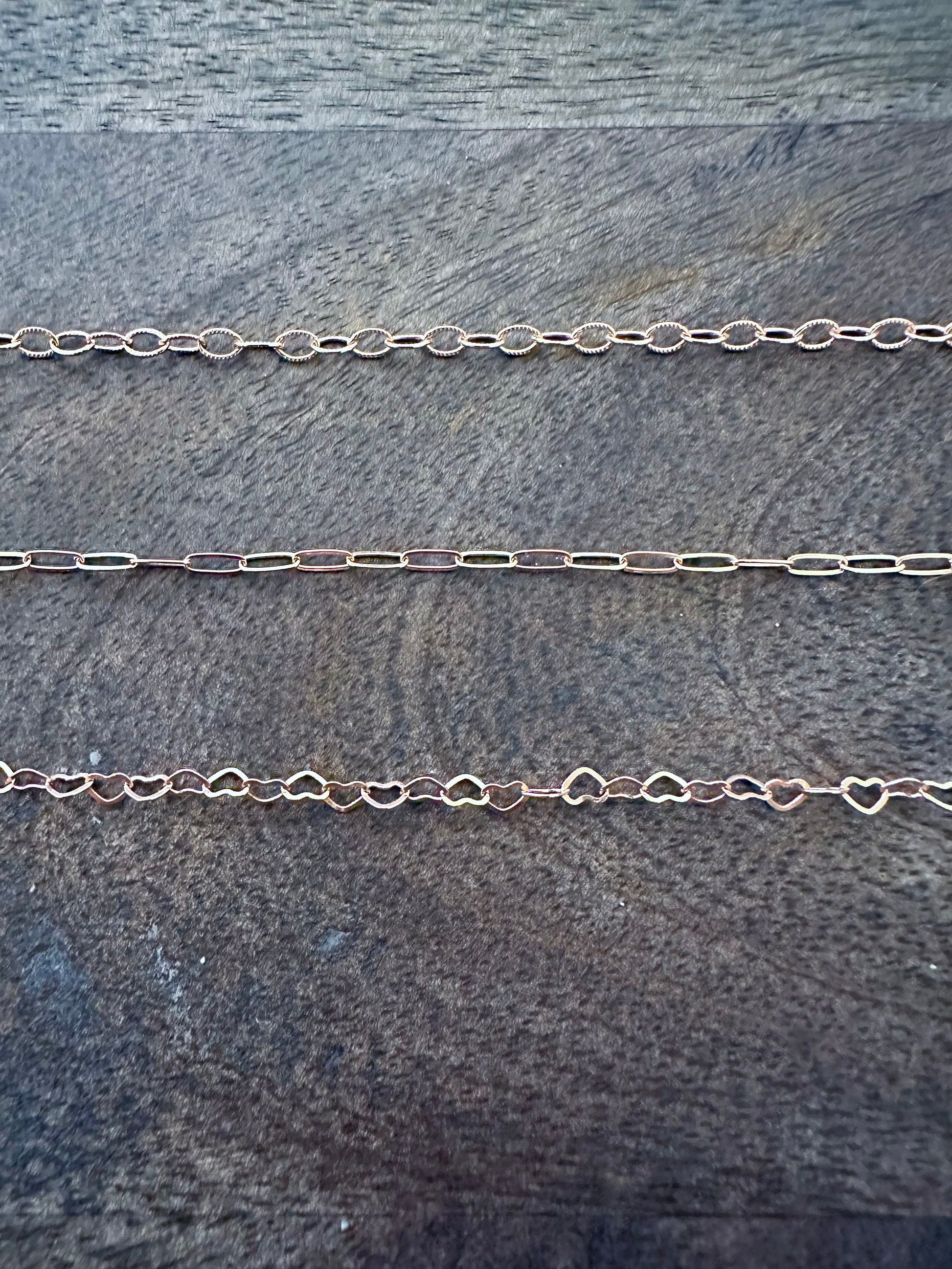 three rose gold chains on a grey wooden backgound. the first chain is alternating textured oval links, the second is a paperclip chain and the thrid are small linked hearts