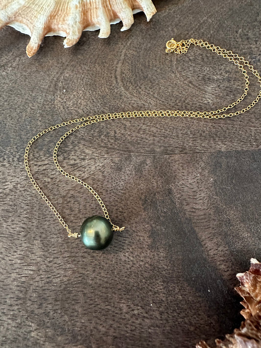 Single black pearl on a gold chain lays on a darekk wooden background with shells in the upper left corner and lower right corner