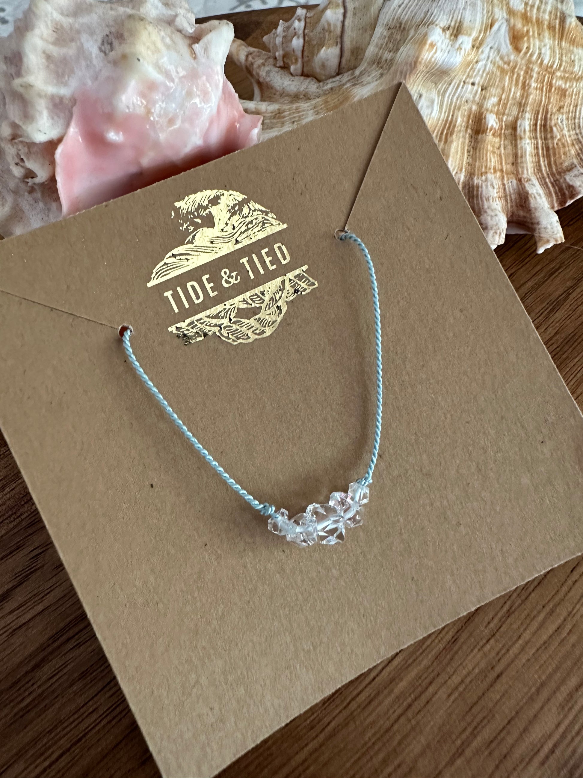 A row of herkimer Diamond stones on a blue silk necklace on a brown craft earring card iwth a gold logo at the top on a wooden background with two shells in the upper edge