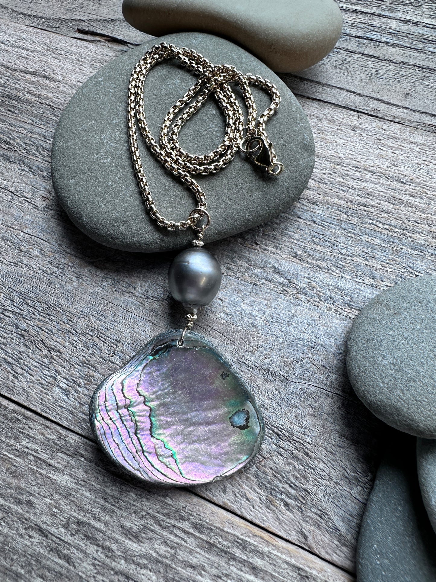 A gold-toned chain necklace with a dark gray pearl bead and a large, irregular iridescent abalone shell pendant, styled on gray river stones over a weathered wood surface.