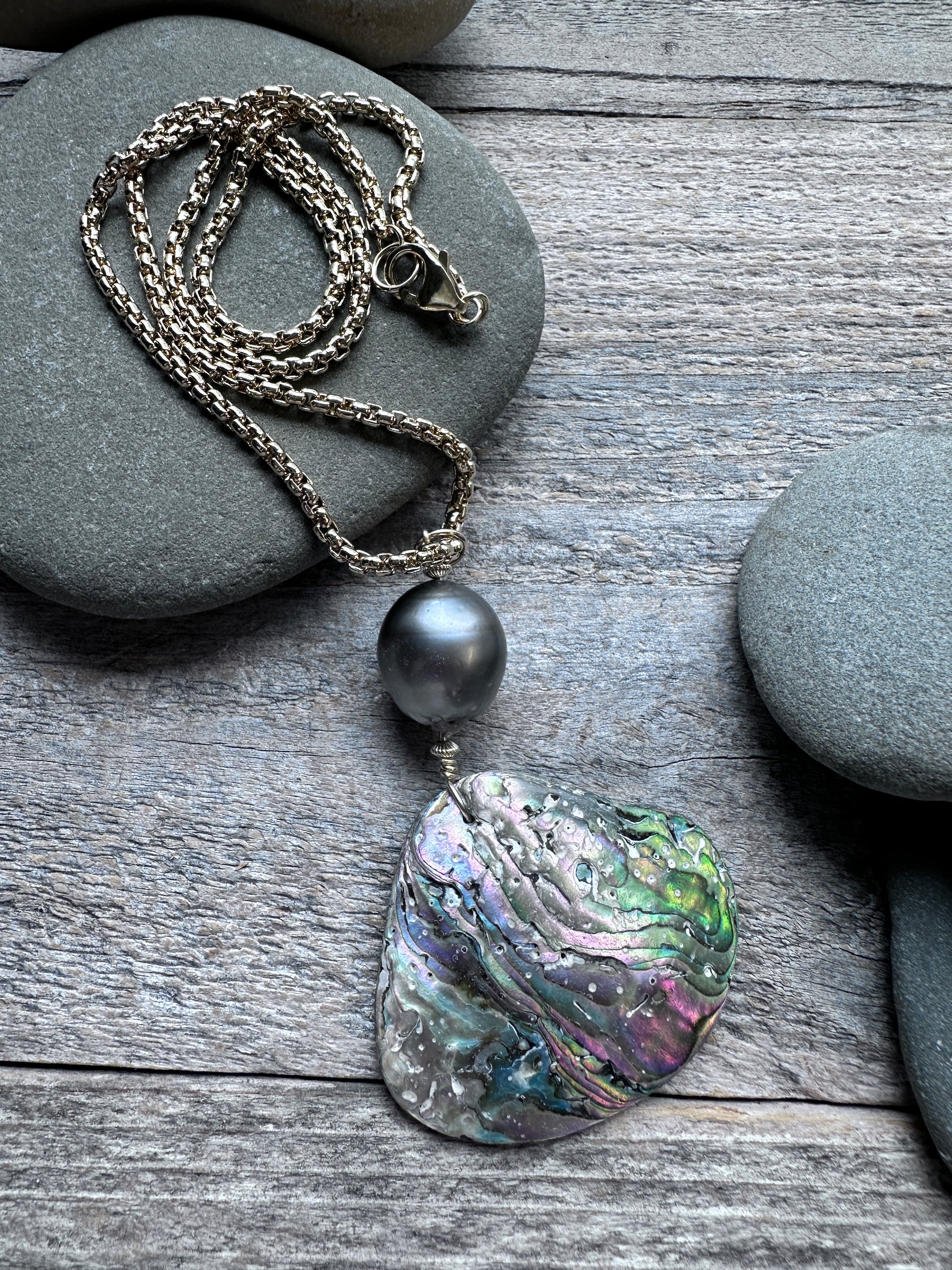 A necklace with a textured, iridescent abalone shell pendant and a dark gray pearl, displayed on a weathered gray wooden surface with smooth gray stones.