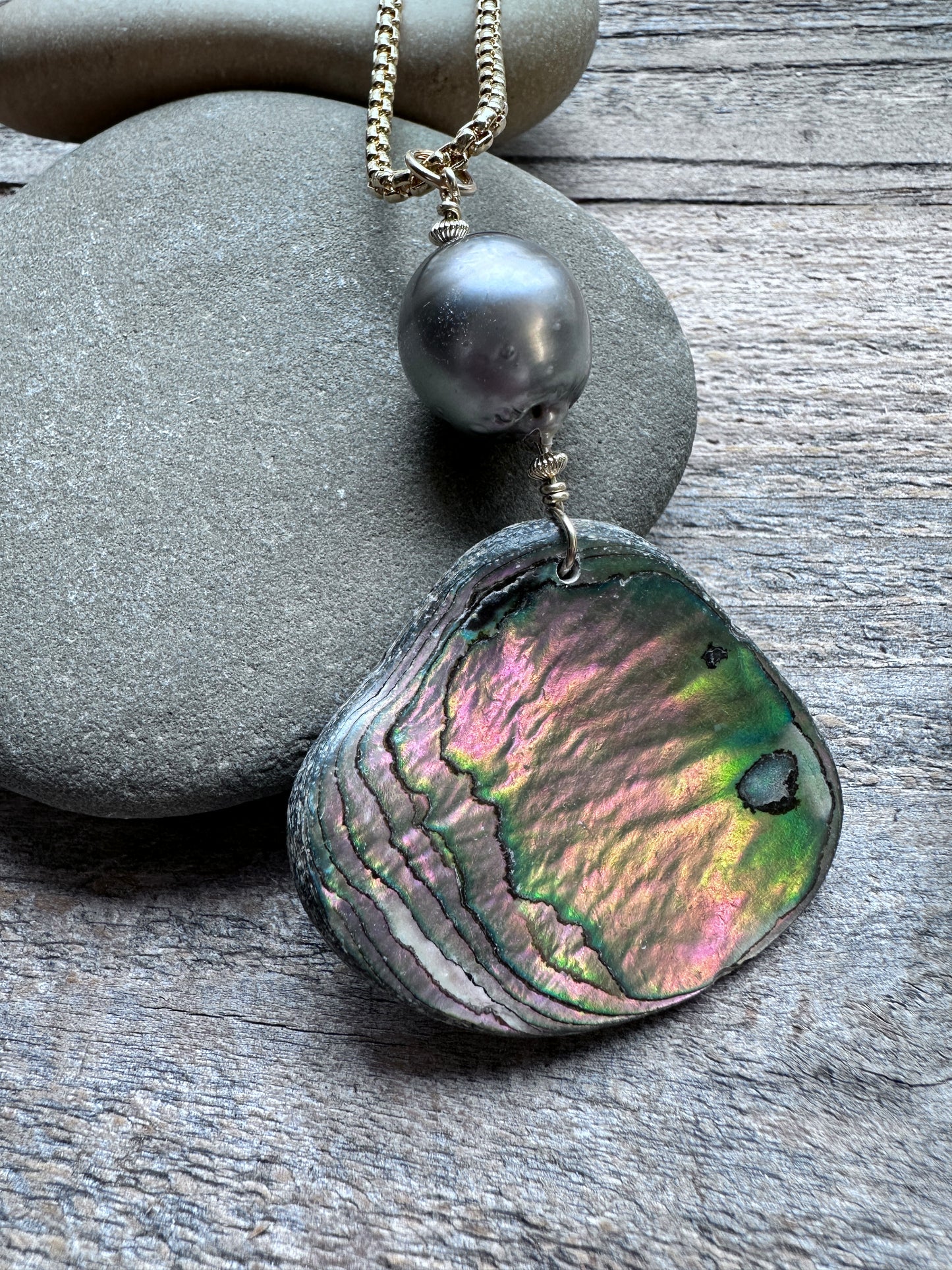 A close-up of a necklace with a large, iridescent abalone shell pendant and a dark gray pearl bead, resting on smooth gray stones over a weathered wood surface.