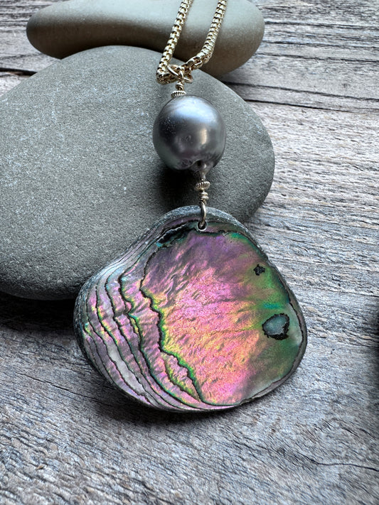 A necklace featuring a large, iridescent abalone shell pendant with pink, green, and blue hues, accented by a dark gray pearl and a gold-toned chain, displayed on gray stones over a weathered wood surface.