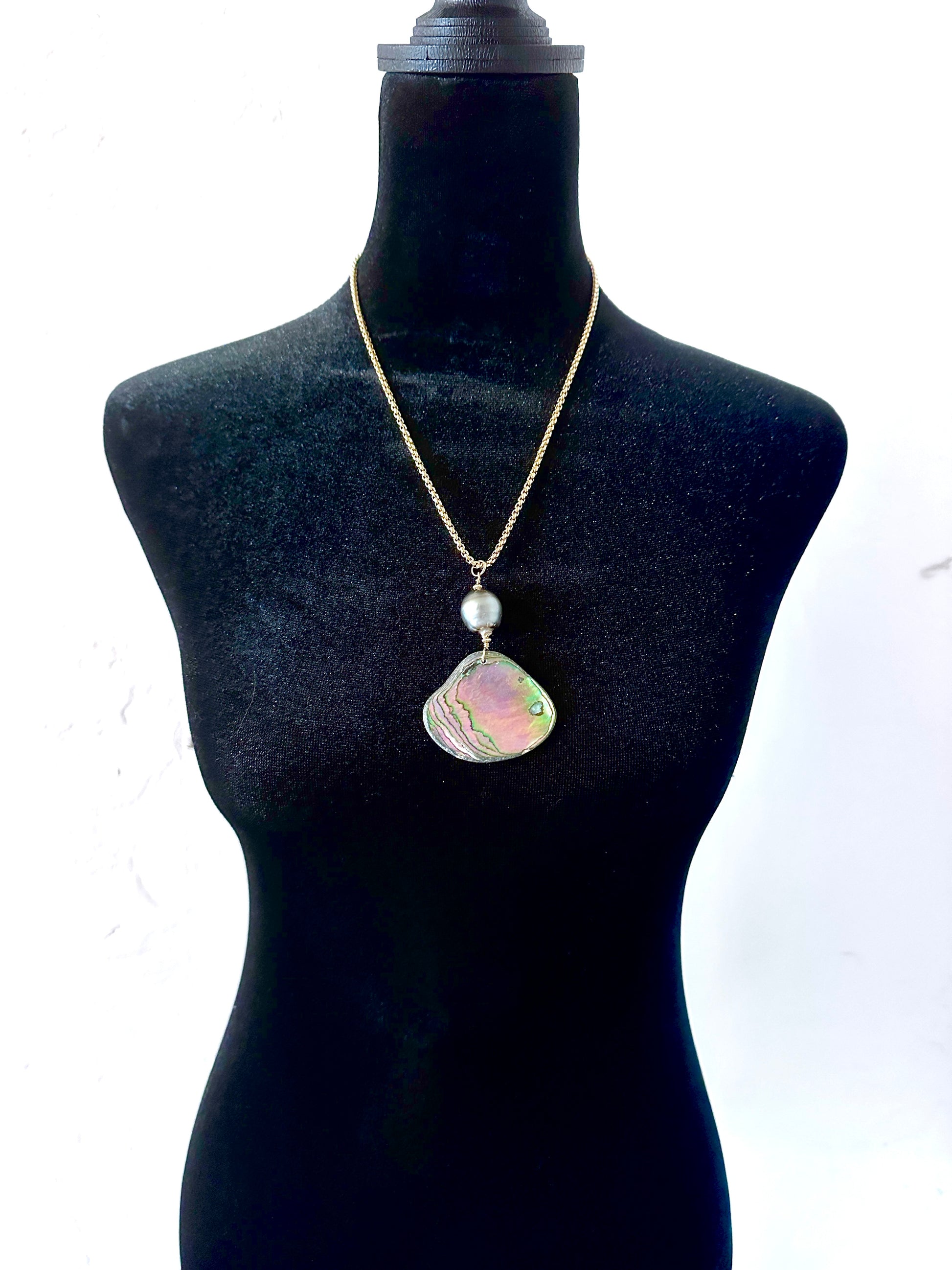 A gold chain necklace with a large, irregularly shaped iridescent shell pendant and a small white pearl, displayed on a black mannequin torso against a white background.