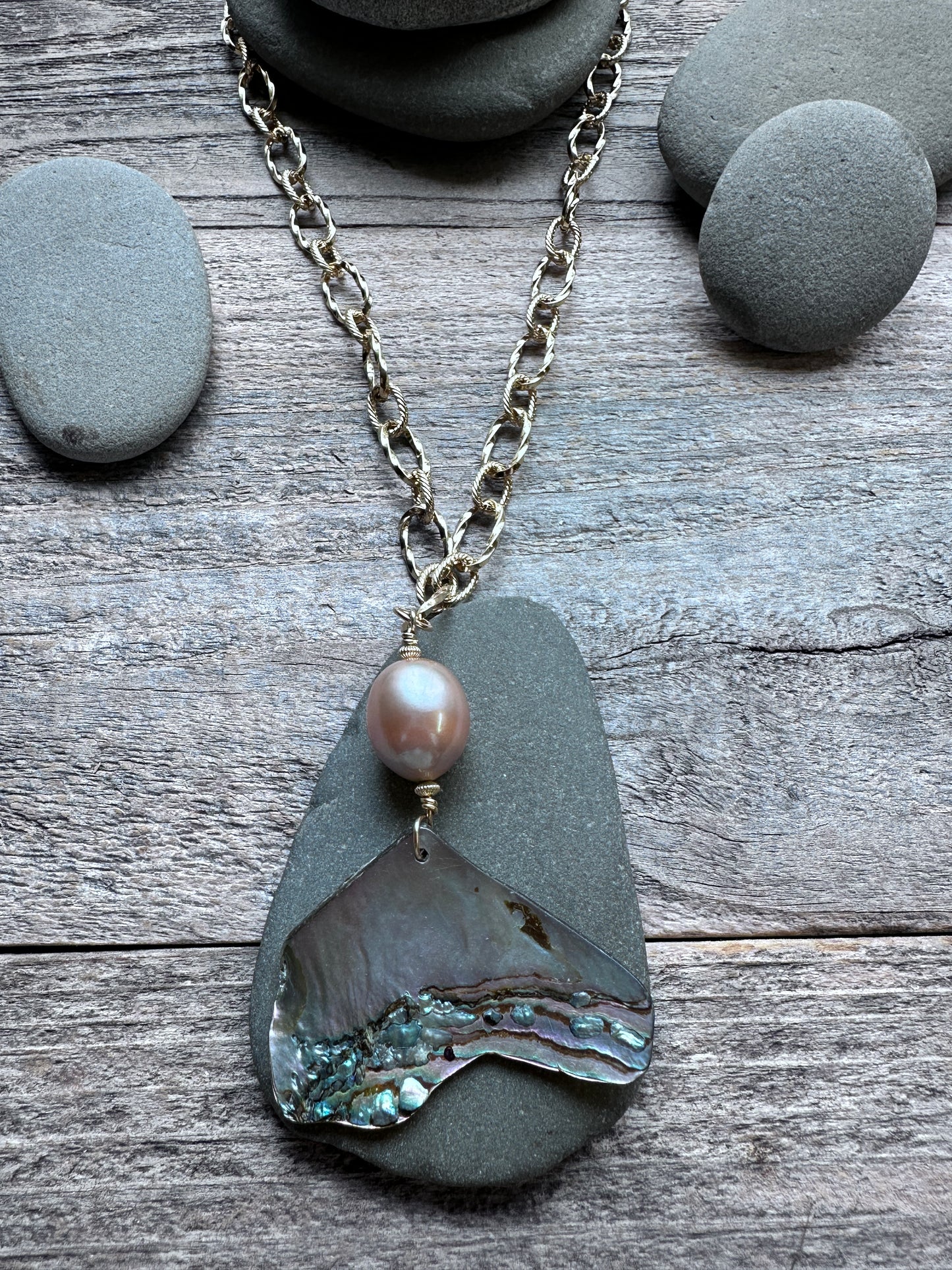 A necklace featuring a large irregular dark gray stone pendant with a polished iridescent abalone shell, a small round pink pearl bead, and an antique silver chain, displayed on a rustic wood surface with smooth gray stones.