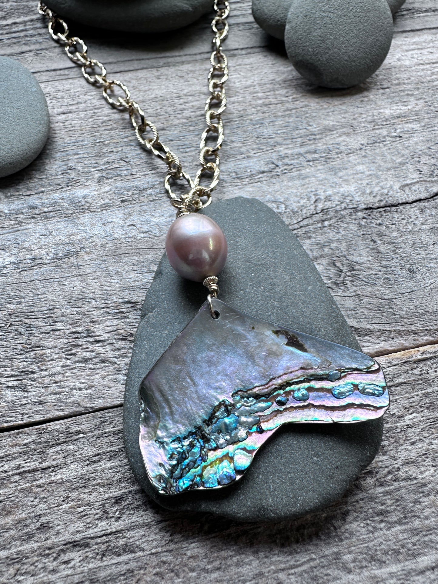 a pink pearl and abalone shell pendant on a large gold colored chain on a grey pebble on a weathered grey background