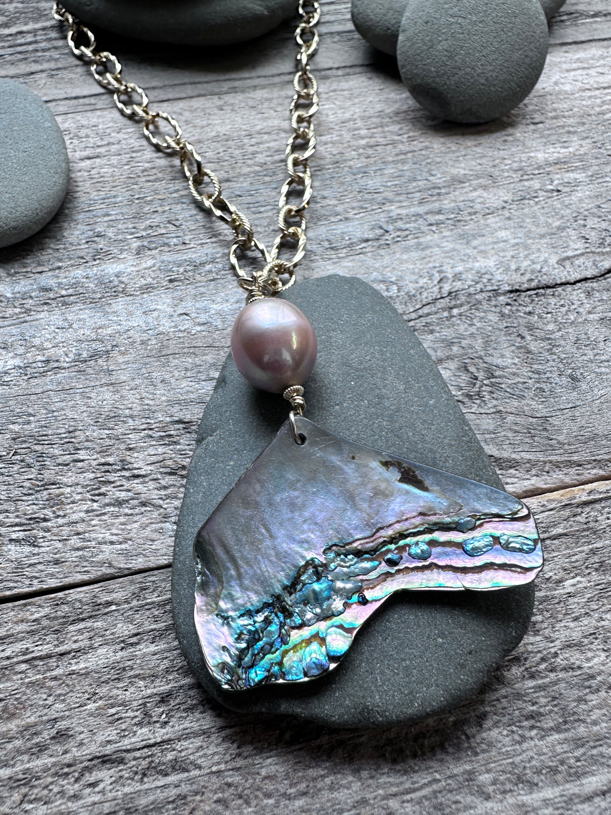 a pink pearl and abalone shell pendant on a large gold colored chain on a grey pebble on a weathered grey background