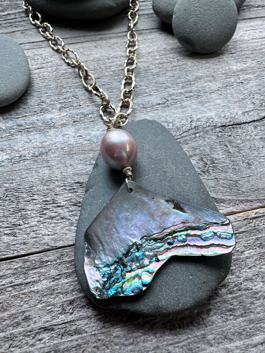 a pink pearl and abalone shell pendant on a large gold colored chain on a grey pebble on a weathered grey background