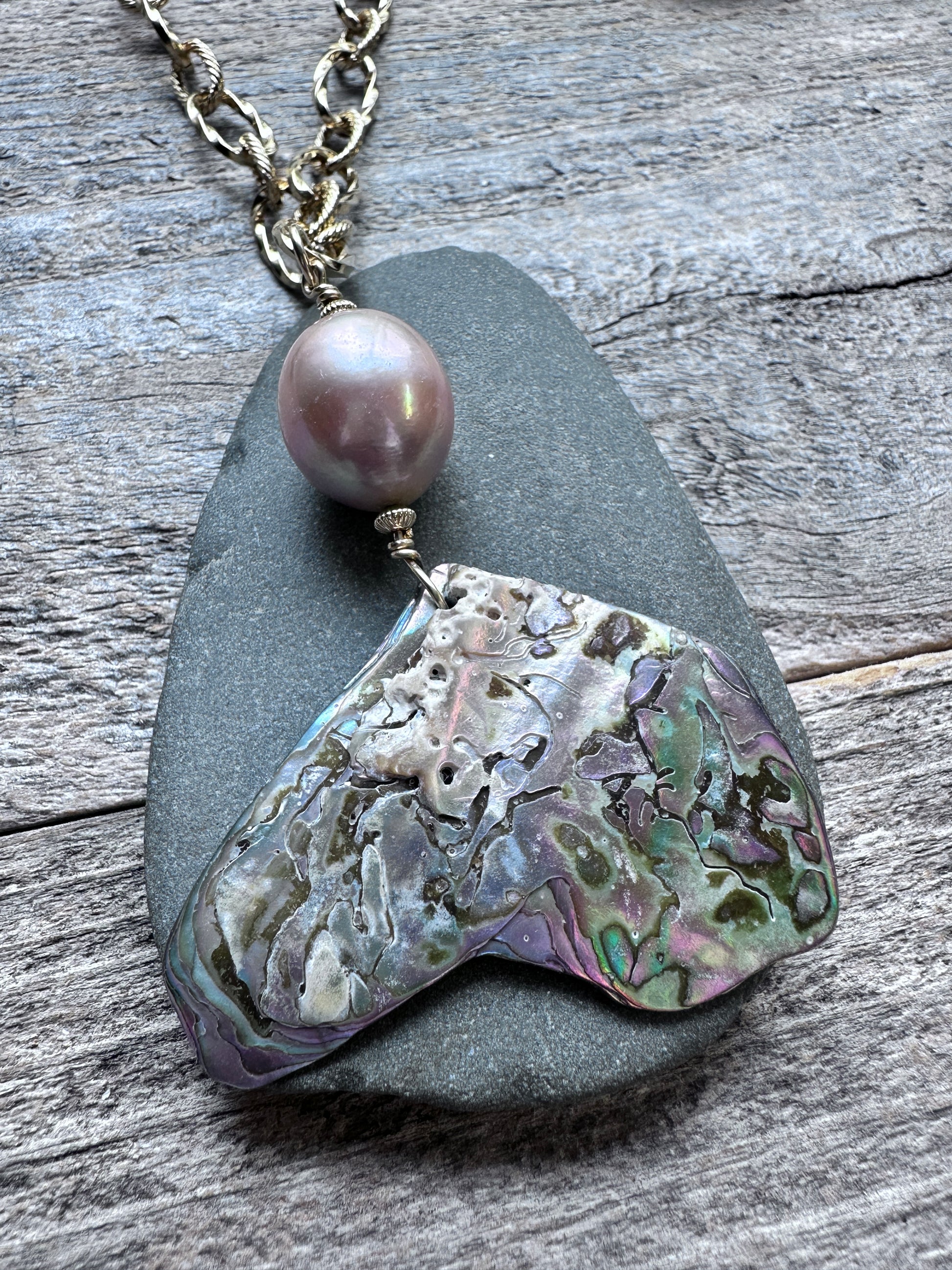 A necklace featuring a large, organic-shaped abalone shell pendant with iridescent purple, green, and silver patterns, suspended from a silver-toned chain and accented by a single round pink pearl, displayed on a dark slate stone over a weathered gray wood background.