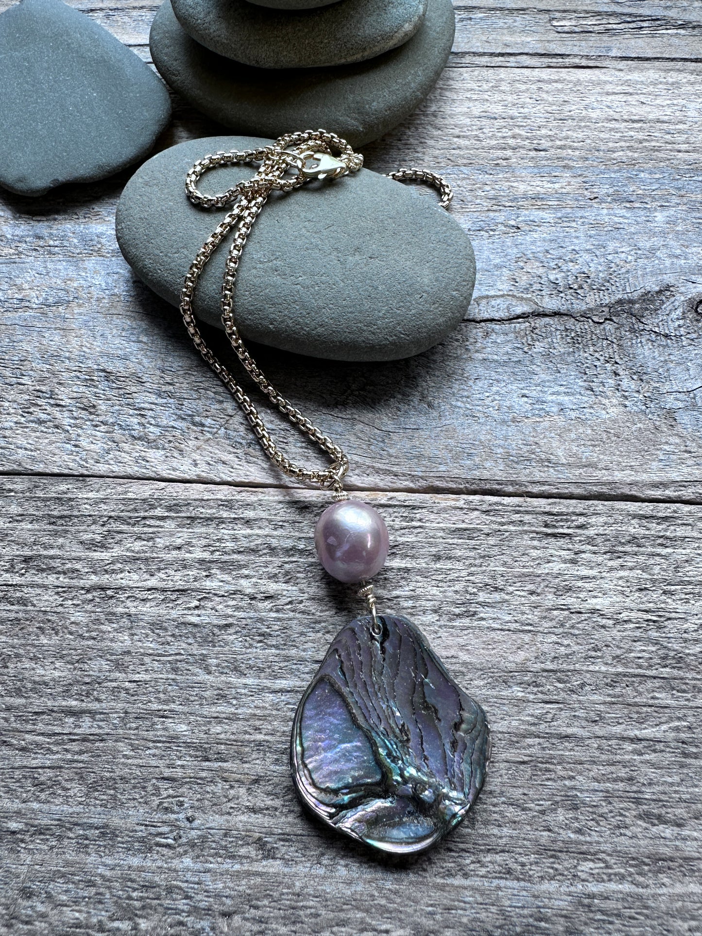 A bohemian-style necklace featuring a large, textured abalone shell pendant and a single light purple pearl, displayed on a rustic gray wooden surface with stacked stones.