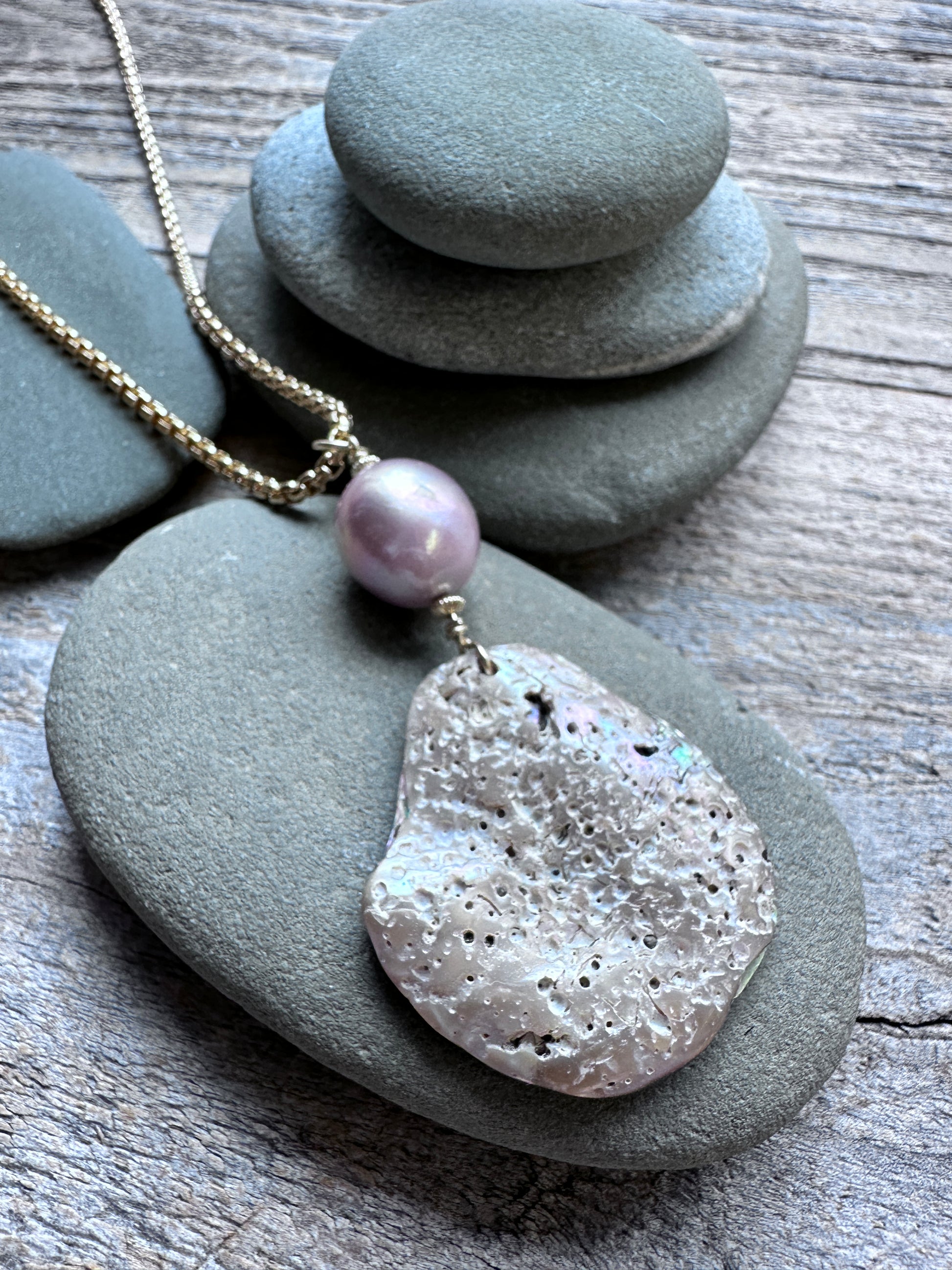A handmade pendant necklace featuring a gold chain, a light purple pearl bead, and a large, textured, abalone pendant, styled on gray river stones over a weathered wood background.