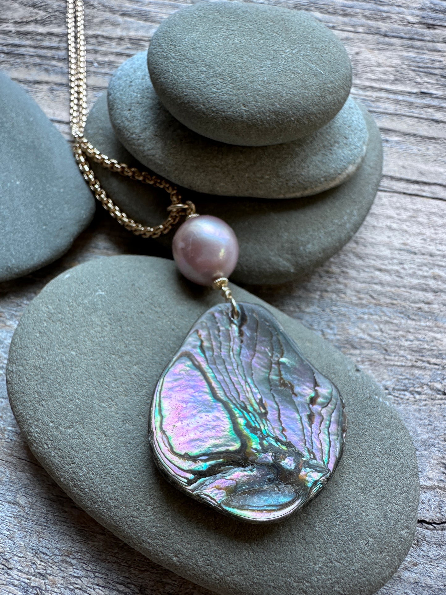 A gold-tone necklace with a teardrop-shaped abalone shell pendant and a single pink pearl bead, styled on stacked gray river stones over a weathered wood surface.