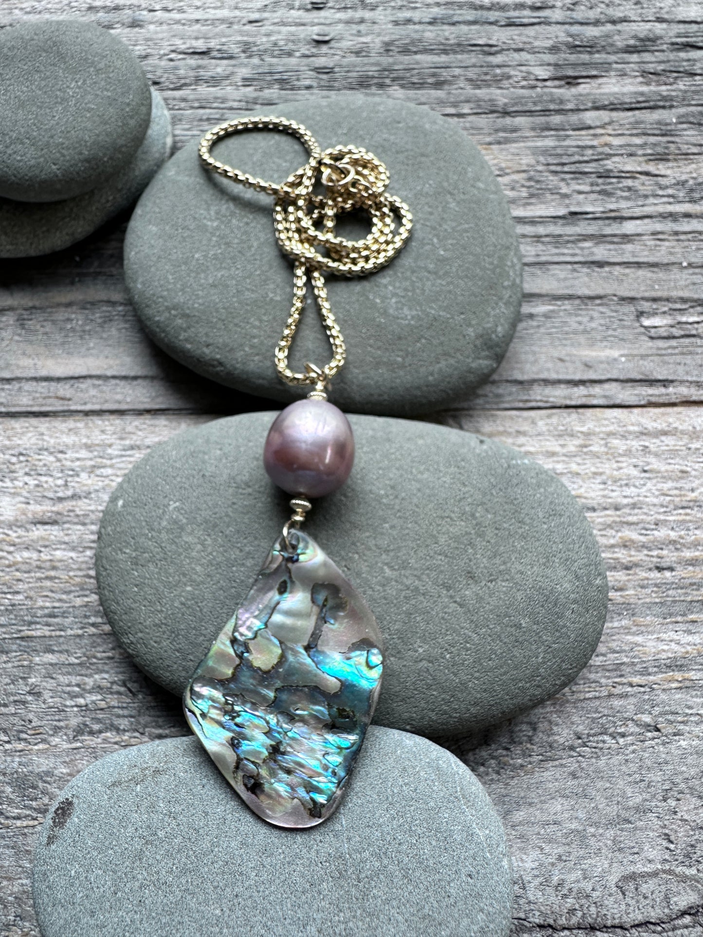 A gold-tone necklace featuring a large teardrop-shaped iridescent abalone shell pendant and a single round purple pearl, displayed on stacked gray river stones over a weathered wood surface.