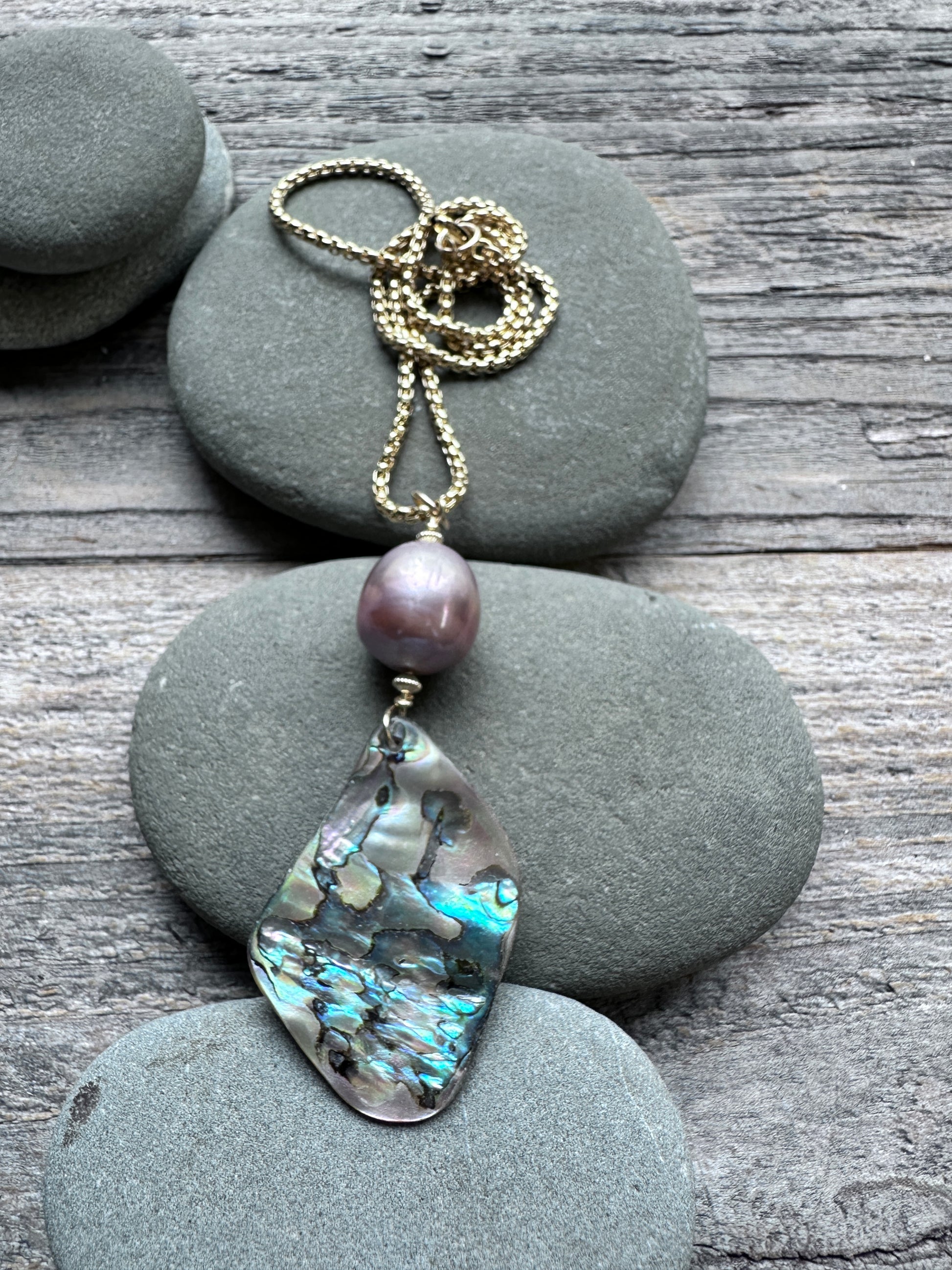 A gold-tone necklace featuring a large teardrop-shaped iridescent abalone shell pendant and a single round purple pearl, displayed on stacked gray river stones over a weathered wood surface.