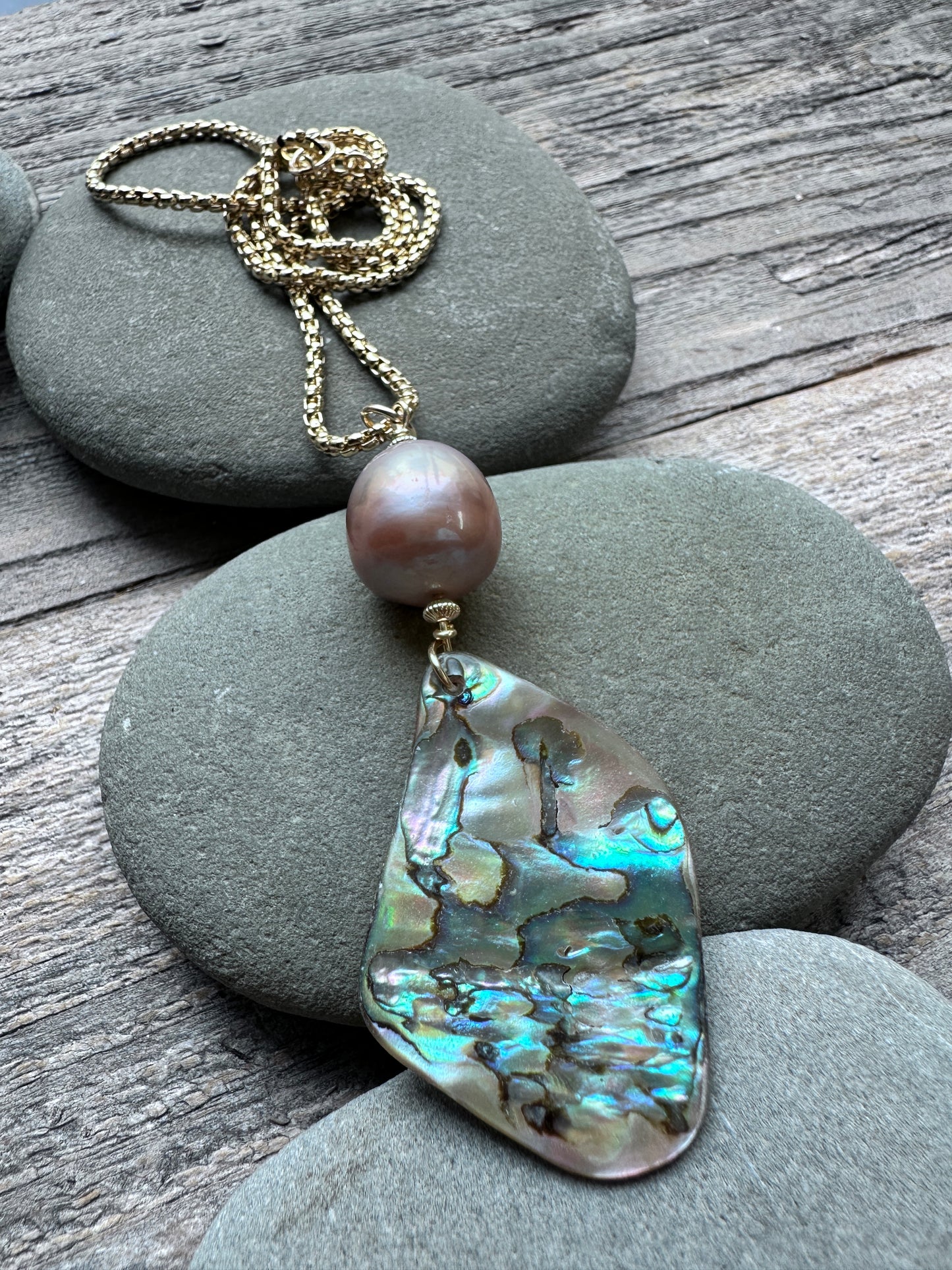 A gold-tone chain necklace featuring a single baroque pearl and a large, irregular abalone shell pendant, displayed on gray river stones over a rustic wooden surface.