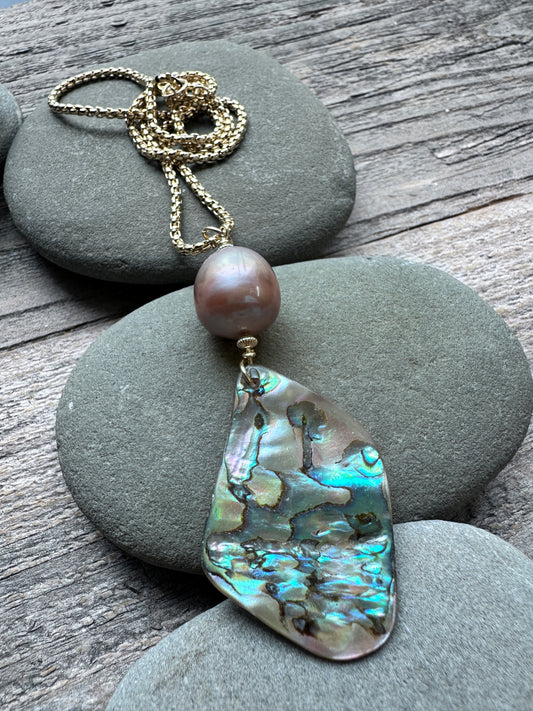 A gold-tone chain necklace featuring a single baroque pearl and a large, irregular abalone shell pendant, displayed on gray river stones over a rustic wooden surface.