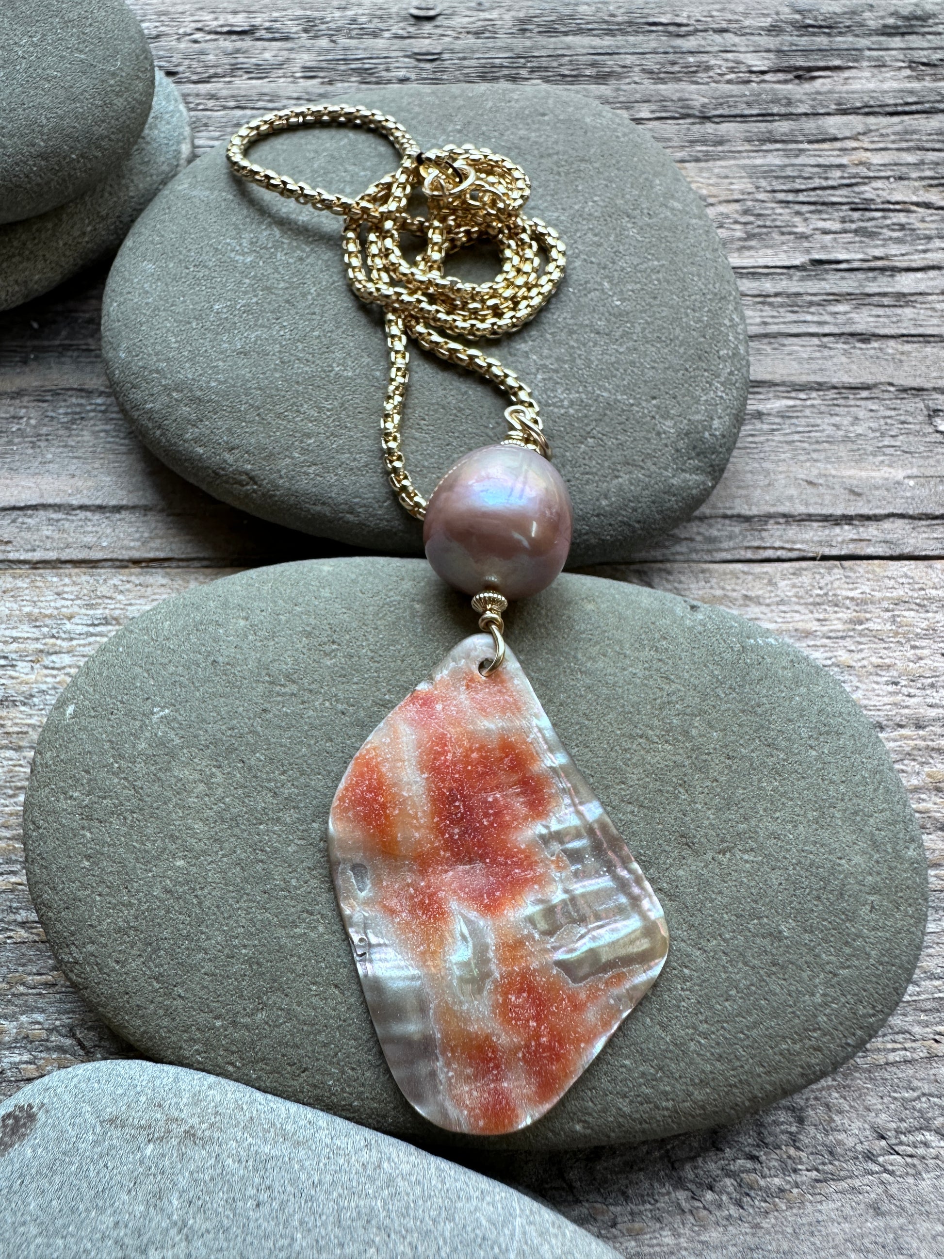 A handmade necklace featuring a gold chain, a single iridescent pearl bead, and a large, irregular polished shell pendant with orange and white marbled patterns, displayed on stacked gray river stones over a rustic wooden surface.