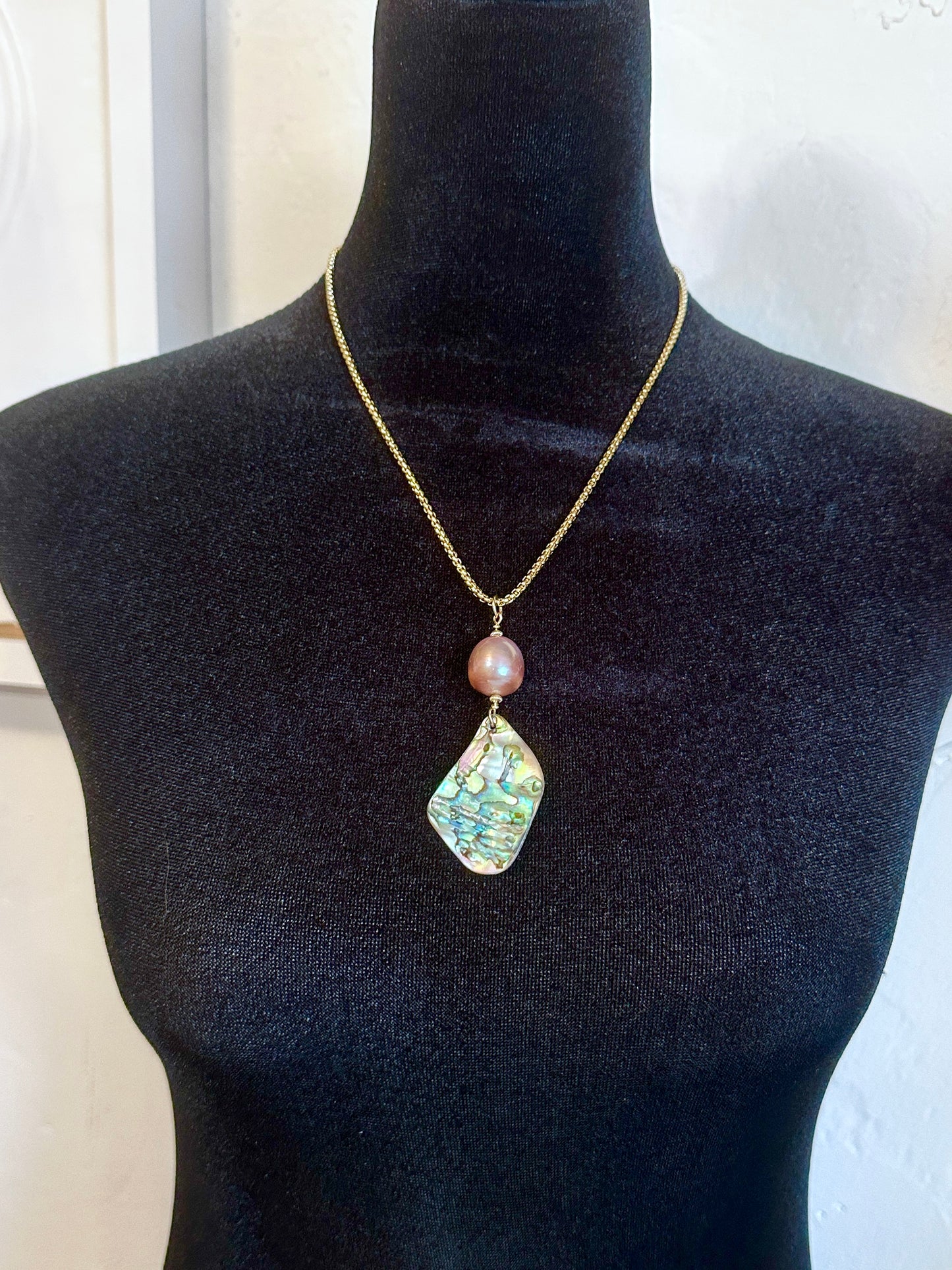 A gold chain necklace with a pink pearl bead and an irregular iridescent green-blue stone pendant, displayed on a black mannequin torso.