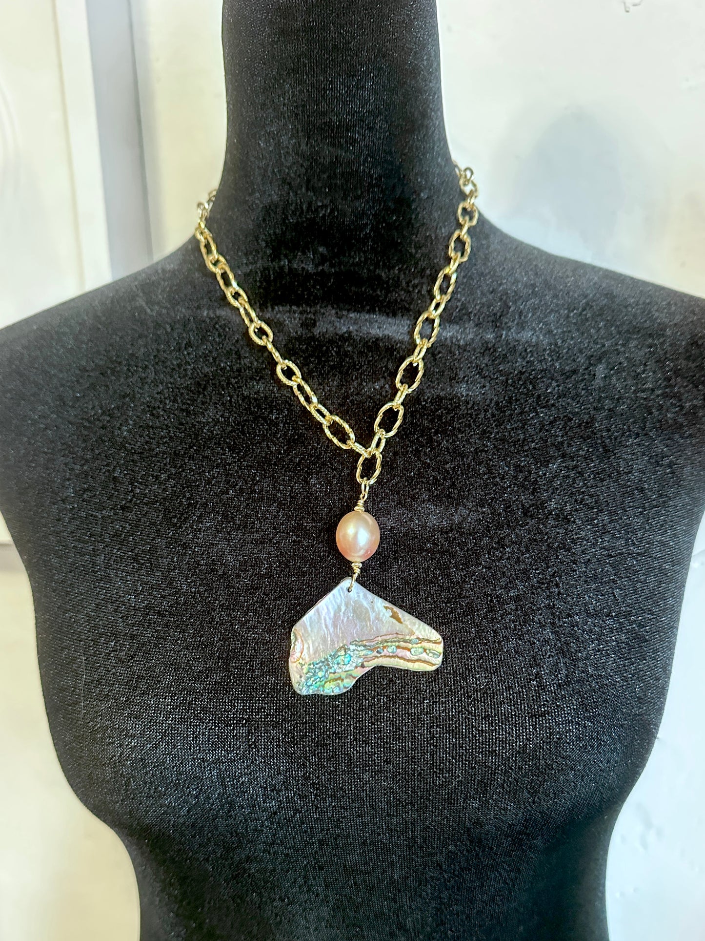 A gold chain necklace displayed on a black mannequin bust, featuring a small iridescent pearl-like bead and a large, irregular shell pendant with shimmering blue, green, and peach tones.