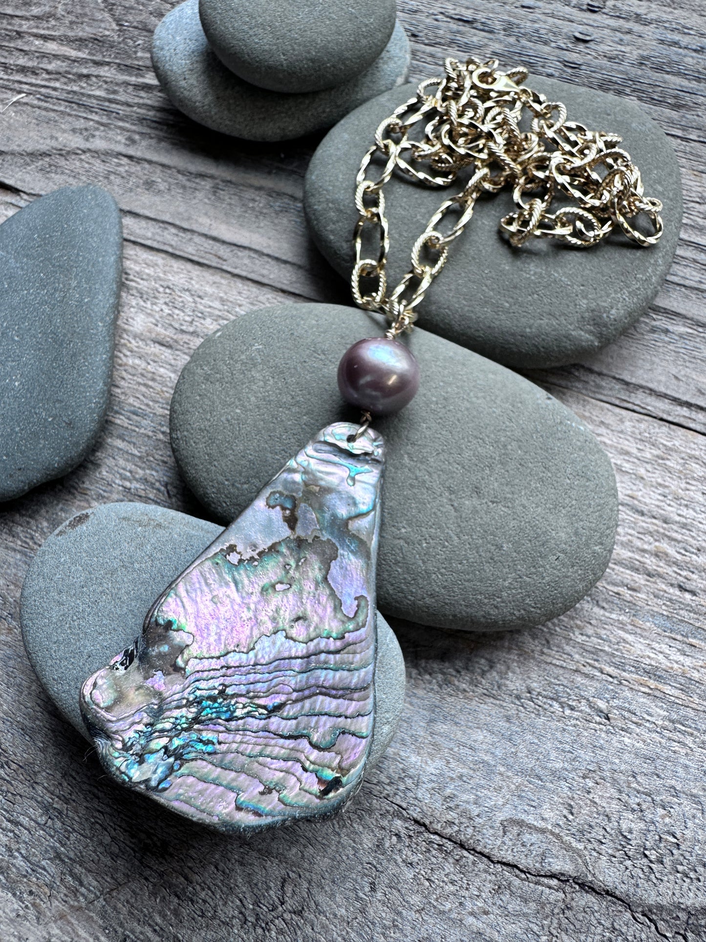 A teardrop-shaped abalone shell pendant necklace with a gold chain and a dark purple pearl accent, displayed on gray river stones over a rustic wooden surface.