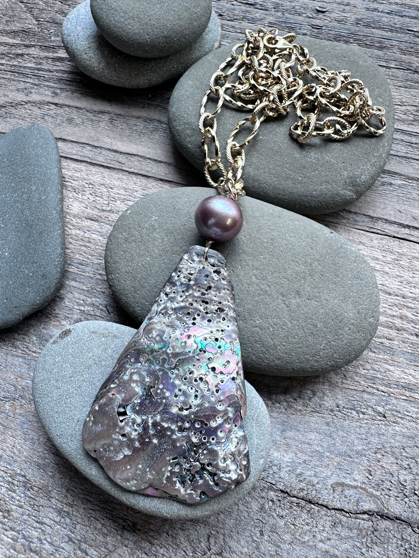 A handmade pendant necklace featuring a large, teardrop-shaped abalone shell pendant with iridescent silver, gray, and purple patterns, a single dark purple pearl bead, and a gold-toned chain, styled on gray river stones over a weathered wood background.