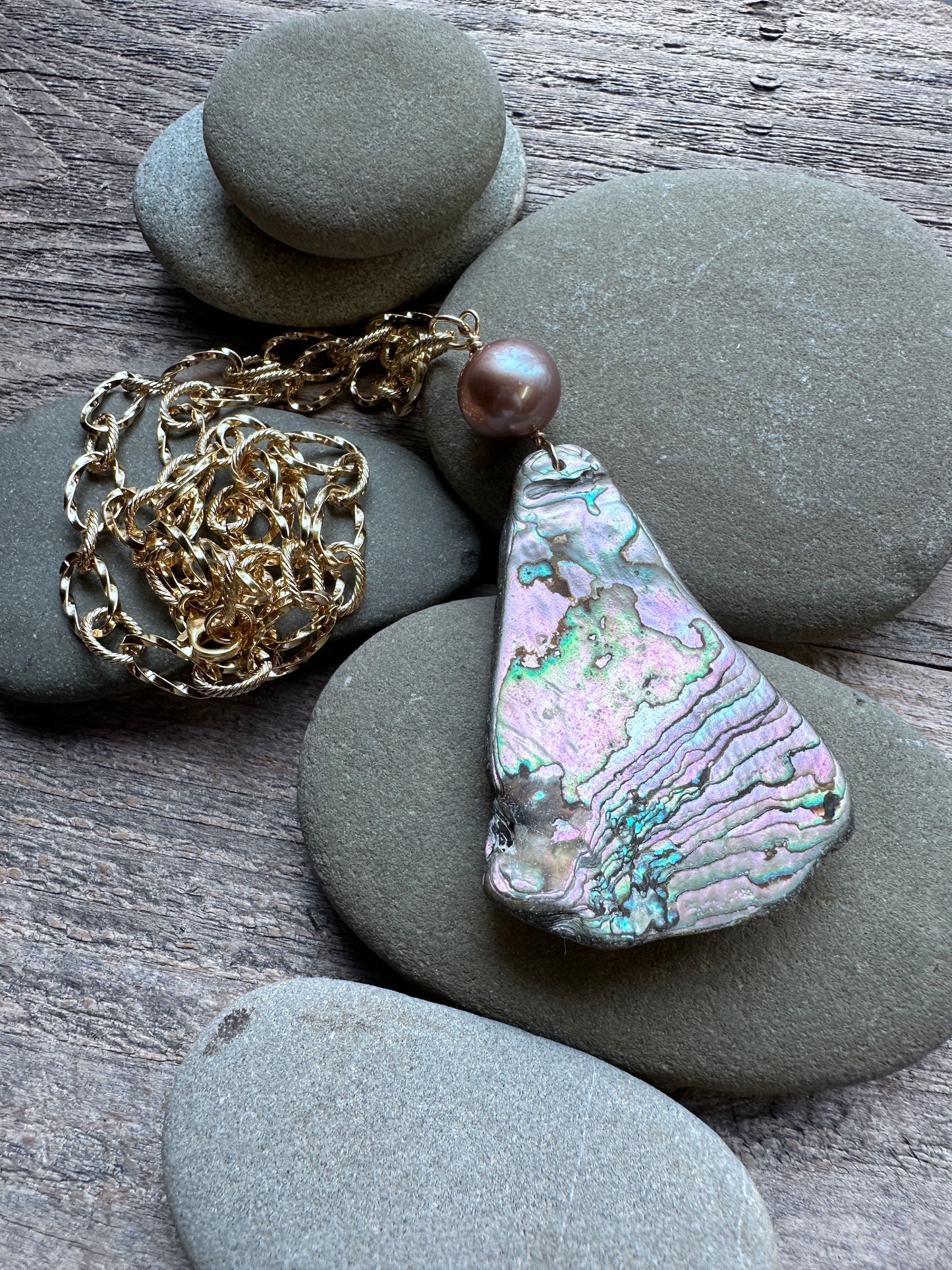 A bohemian-style pendant necklace featuring a teardrop-shaped iridescent abalone shell charm with pink, green, and blue hues, a single round pearl bead, and a chunky gold chain, styled on smooth gray river stones over a rustic wooden surface.