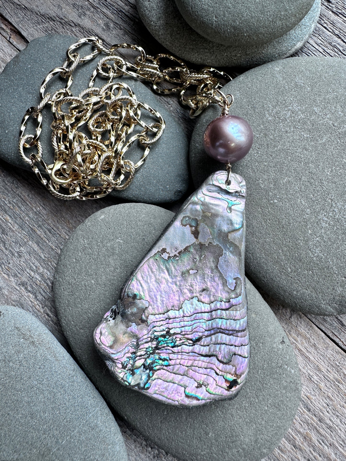 A teardrop-shaped abalone shell pendant necklace with a textured iridescent shell and a single dark purple pearl bead, displayed on smooth gray stones over a wooden surface with a gold chain.