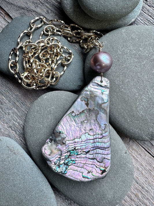A teardrop-shaped abalone shell pendant necklace with a textured iridescent shell and a single dark purple pearl bead, displayed on smooth gray stones over a wooden surface with a gold chain.