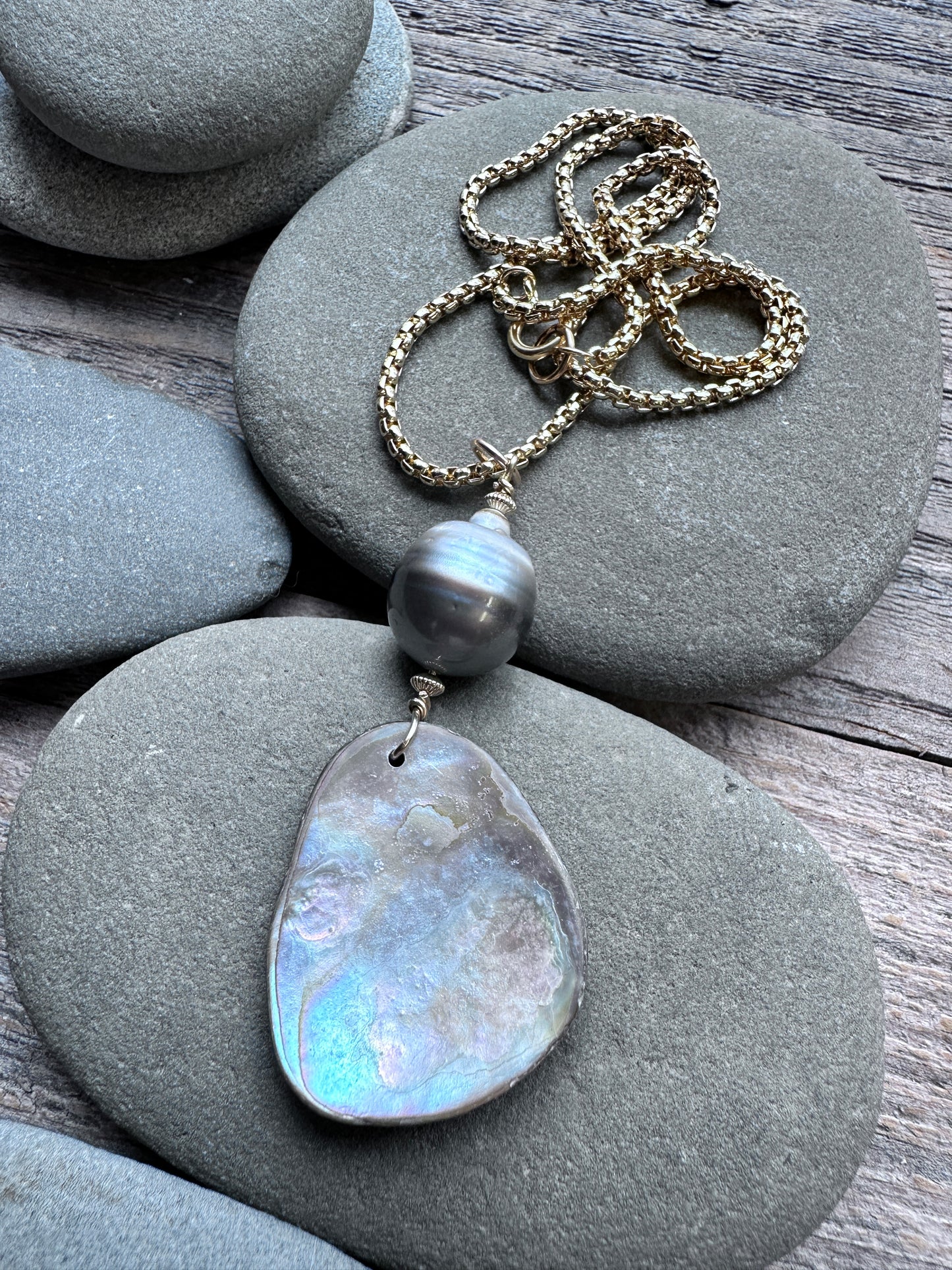A gold-tone chain necklace with a large, irregular iridescent shell pendant and a single dark gray pearl bead, displayed on smooth gray river stones over weathered wood.