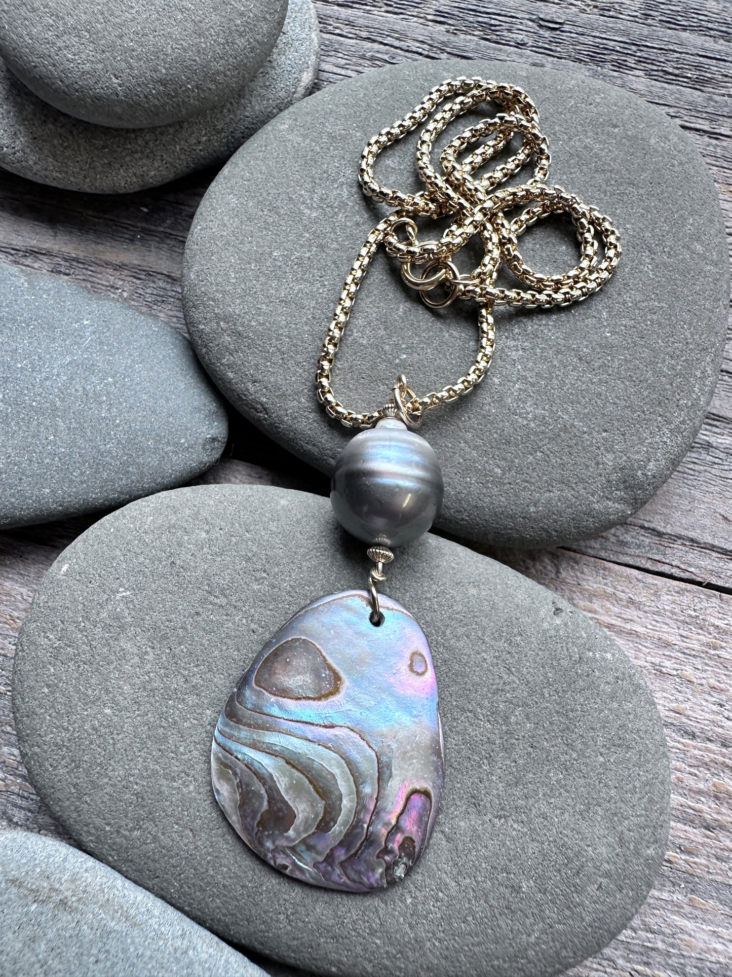 A necklace with a gold chain, a dark gray pearl bead, and a large iridescent abalone shell pendant, displayed on smooth gray stones over a wooden surface.