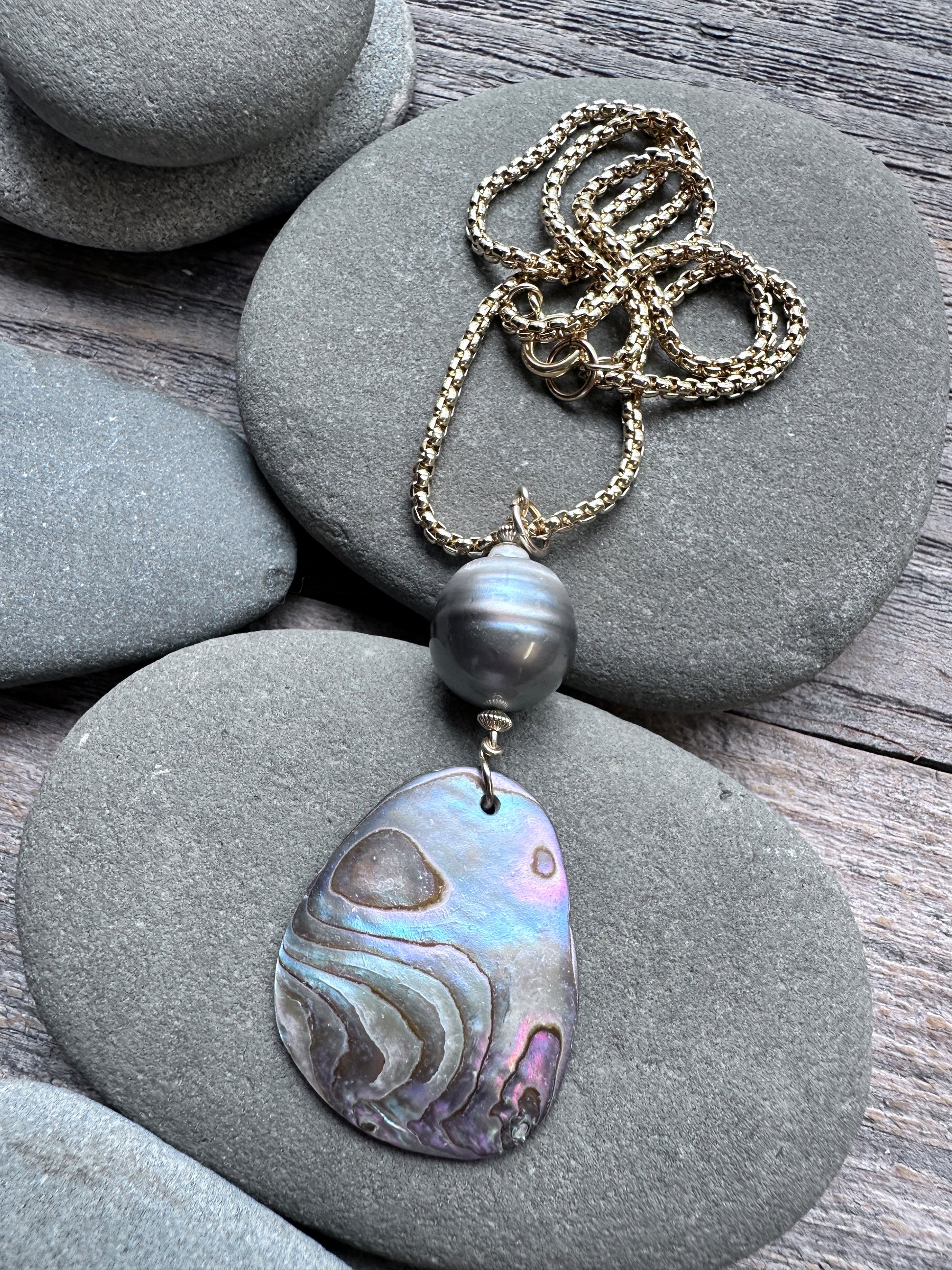 A necklace with a gold chain, a dark gray pearl bead, and a large iridescent abalone shell pendant, displayed on smooth gray stones over a wooden surface.