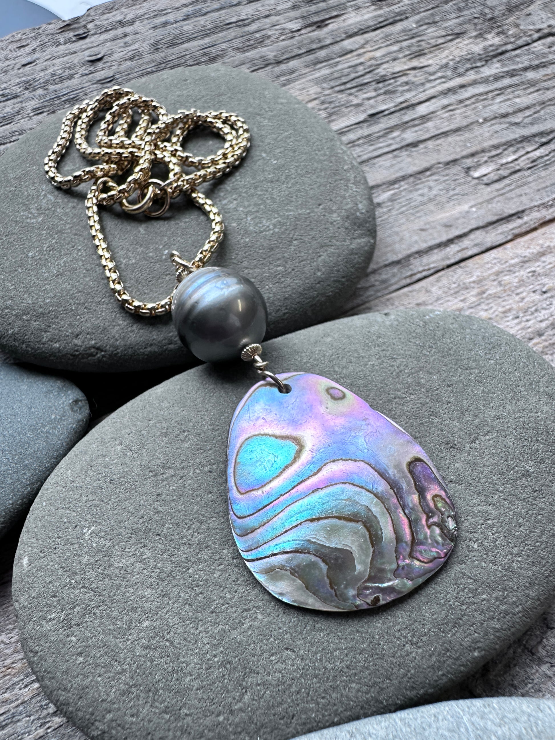 A teardrop-shaped abalone shell pendant necklace with iridescent blue, purple, and silver patterns, hanging from a gold chain with a dark gray bead, displayed on smooth gray stones over a weathered wooden surface.