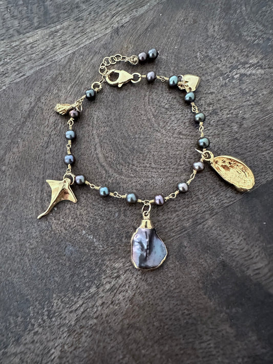 a charm bracelet with gold ocean themed charms on a grey wooden background. a large dark purpleish black odd shaped pearl is in the center.