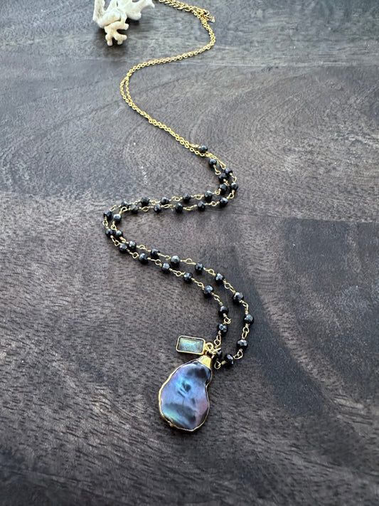 a necklace with black rosary beads and a purpleish black odd shaped pearl with a small rectangular labradorite bead on a grey  wooden background. a small white piece of coral is in the upper left corner