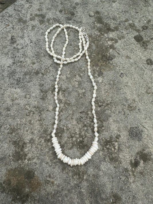 a long neckalce with white little pearls and a bar of white puka shells on a grey stone background. 