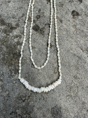 small white seed pearls steung on a necklace with a bar of puka shells in the center on a grey stone background
