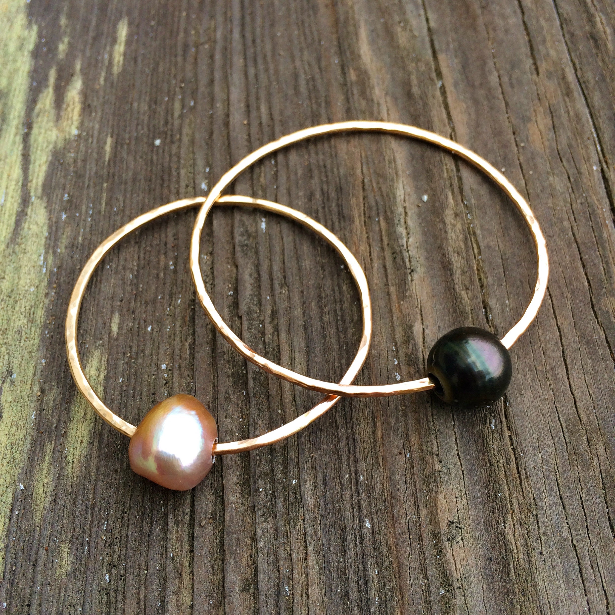 on a dark wood background are two bangles, one with a pink pearl and one with a black pearl.