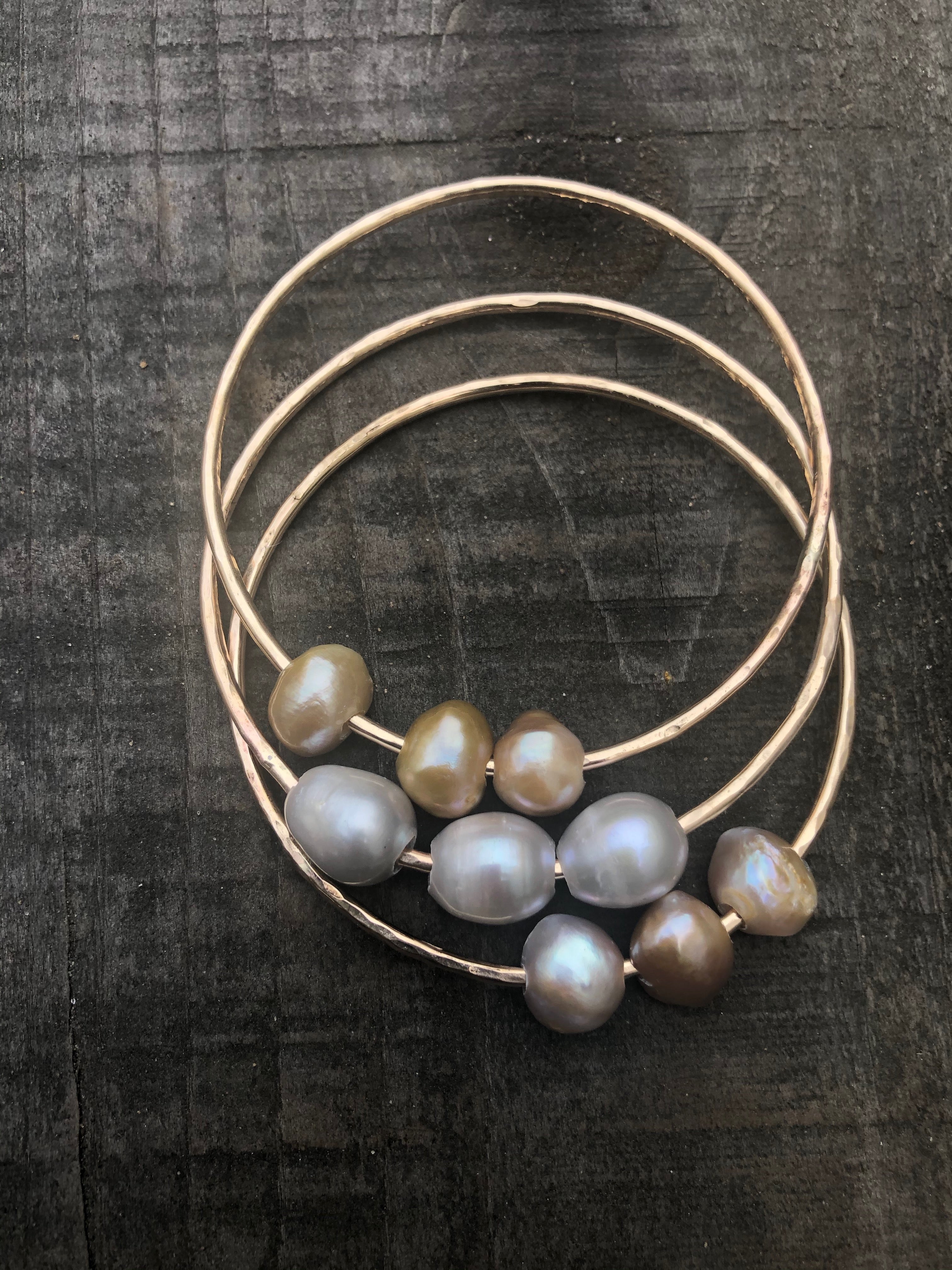 on a dark wood background there are three bangles with three pearls on each. One pink, one white and one golden colored pearls.
