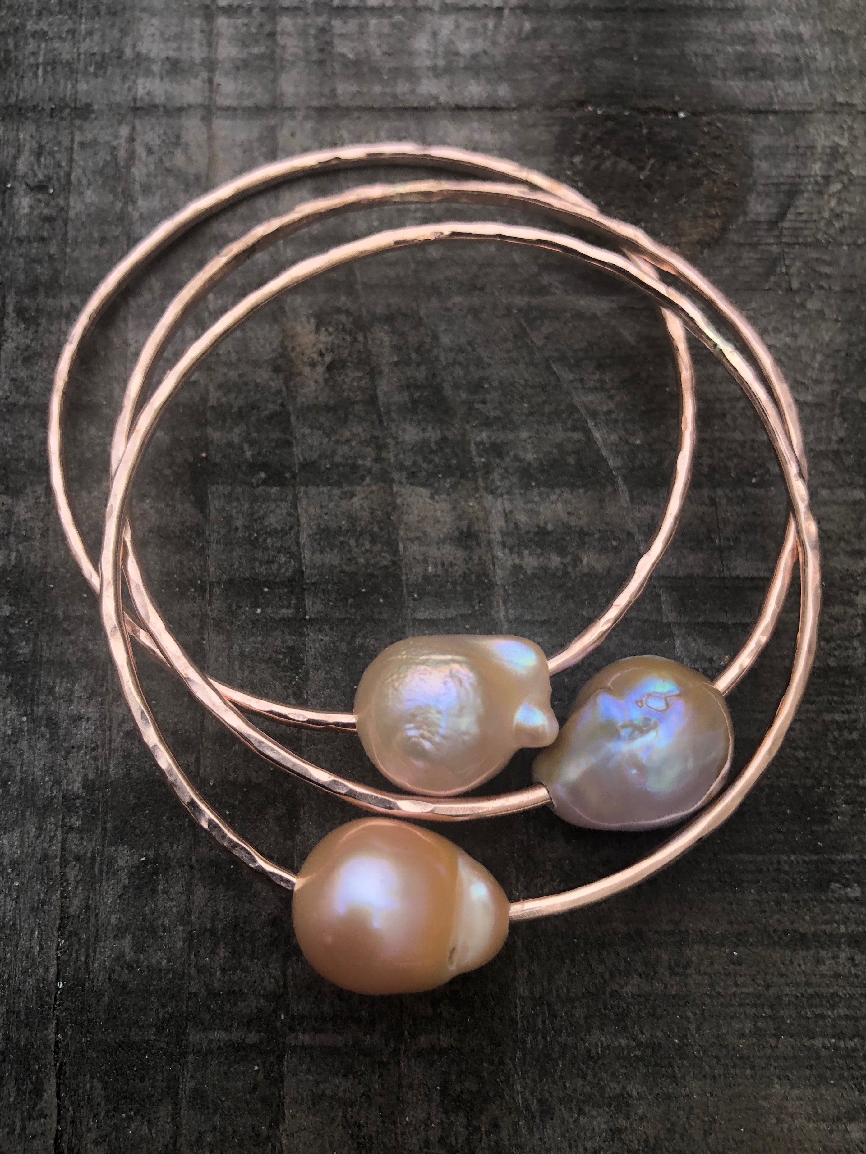 Three gold bangles with large oddly shaped fireball pearls in light hues of pink and peach and white on a wooden background.