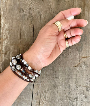 long strand of Tahitian pearls, aquamarine, mini puka shells and grey seed beads  with a dark brown leather tie wrapped around a womans wrist as a bracelet laying on a grey wooden background