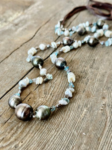 long strand of Tahitian pearls, aquamarine, mini puka shells and grey seed beads  witha dark brown leather tie on a grey wooden background