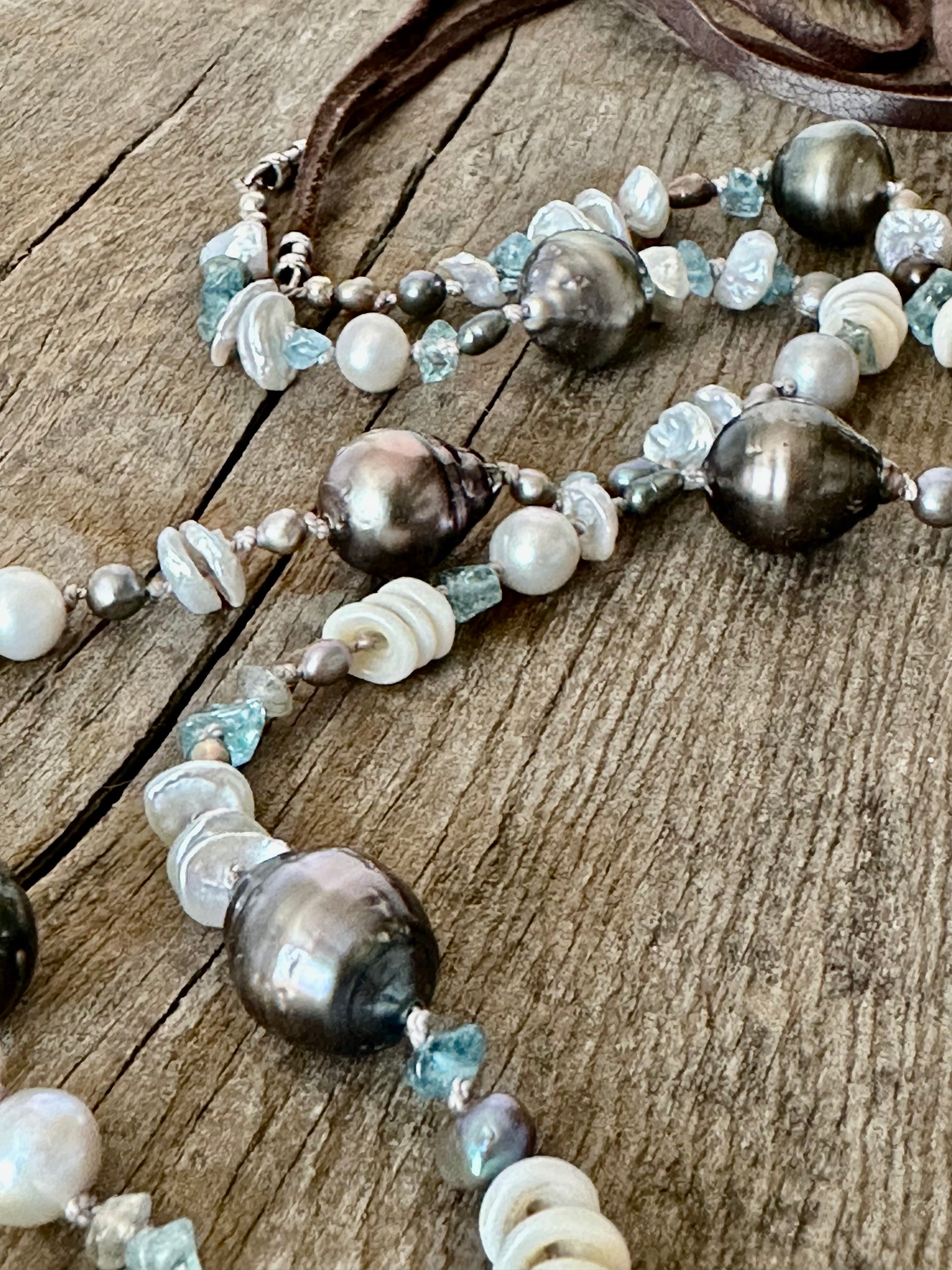 close up of long strand of Tahitian pearls, aquamarine, mini puka shells and grey seed beads  with a dark brown leather tie on a grey wooden background
