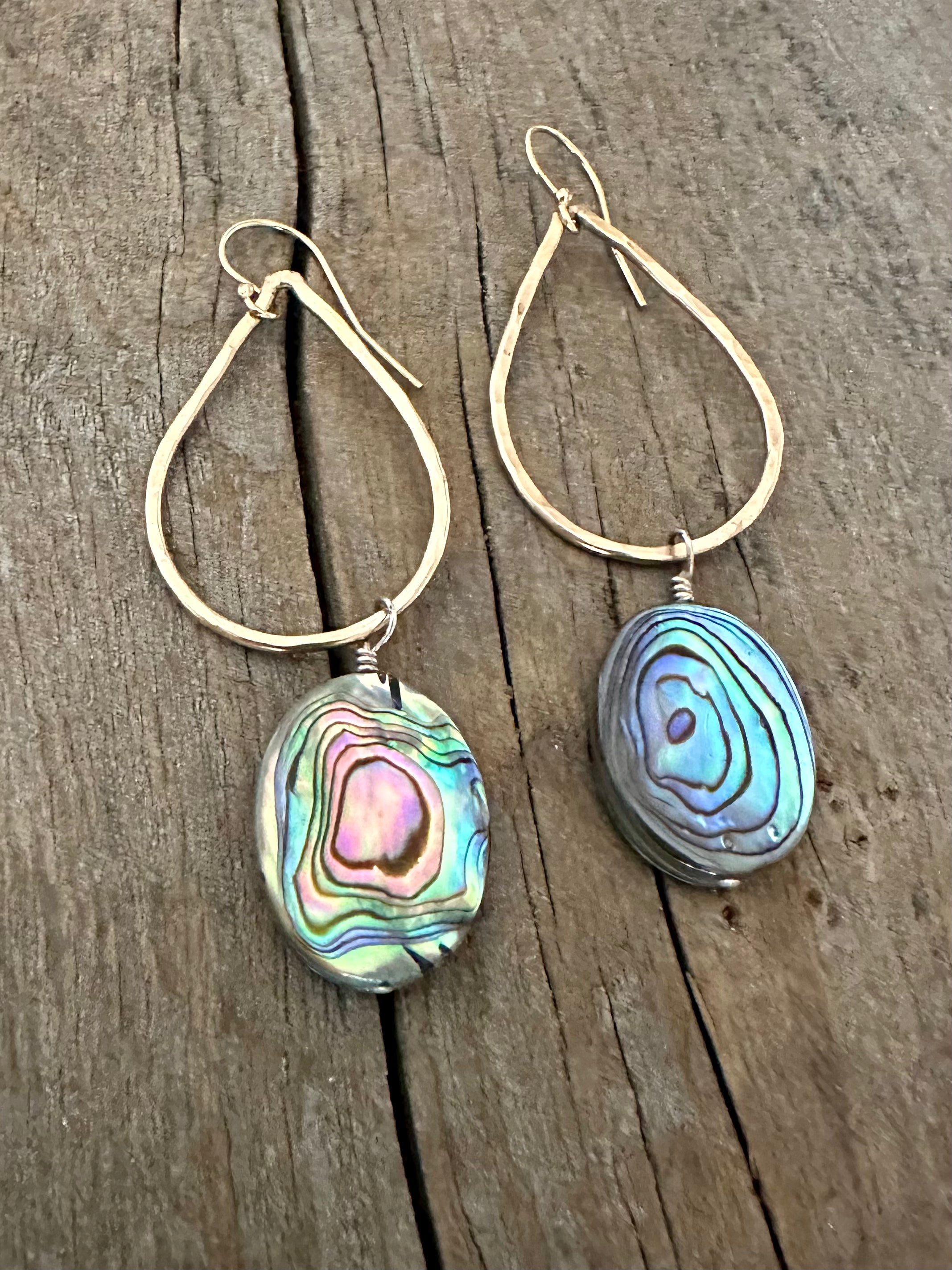 rainbow abalone shell in oval form hanging off of a teardrop shaped earring on a wooden background