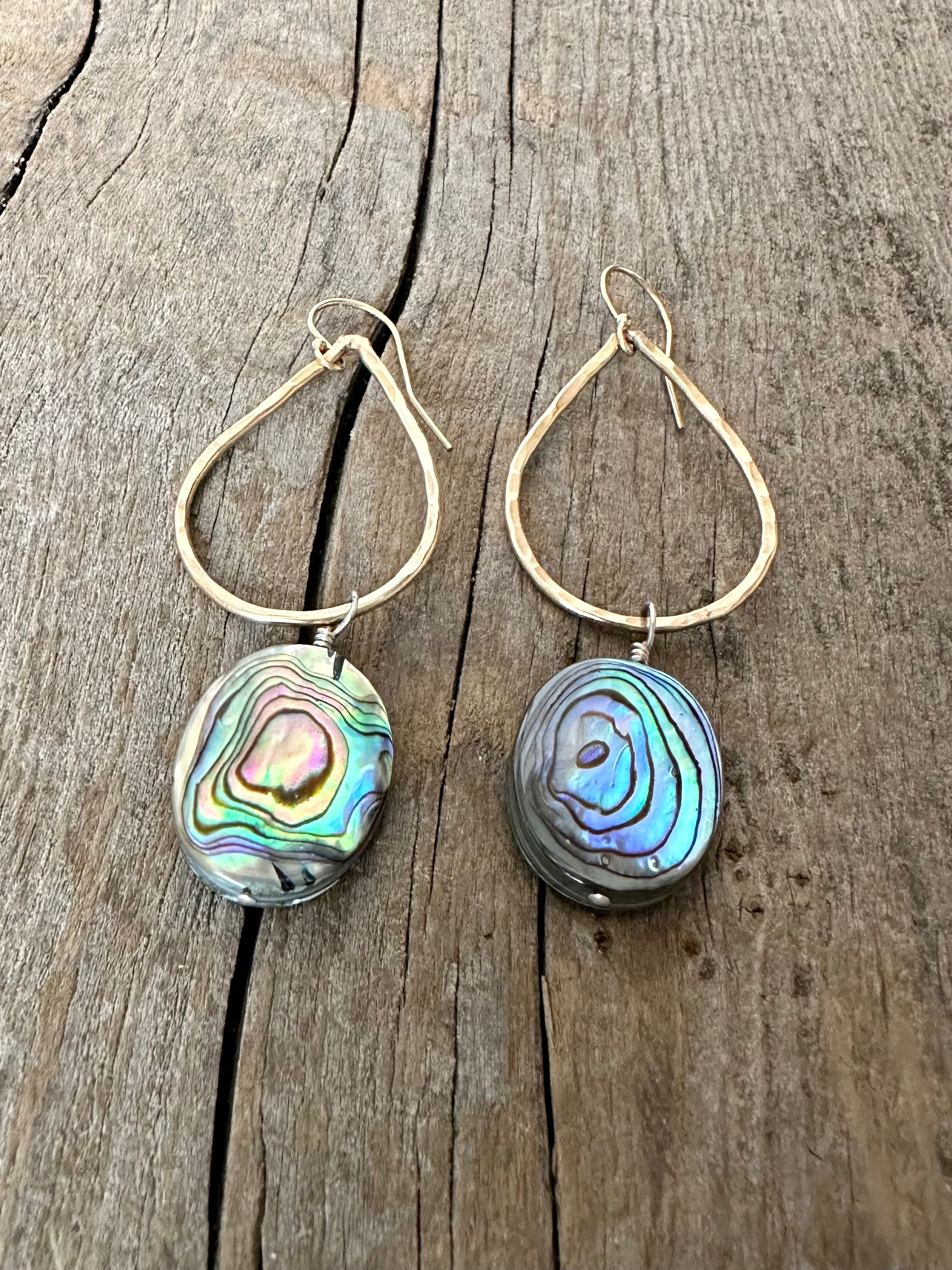 rainbow abalone shell in oval form hanging off of a teardrop shaped earring on a wooden background