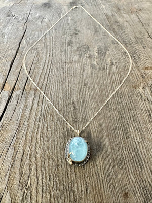 Blue Aquamarine oval stone set in silver with gold prongs and a small gold ball in the lower left corner on a gold chain laying on a wooden background.