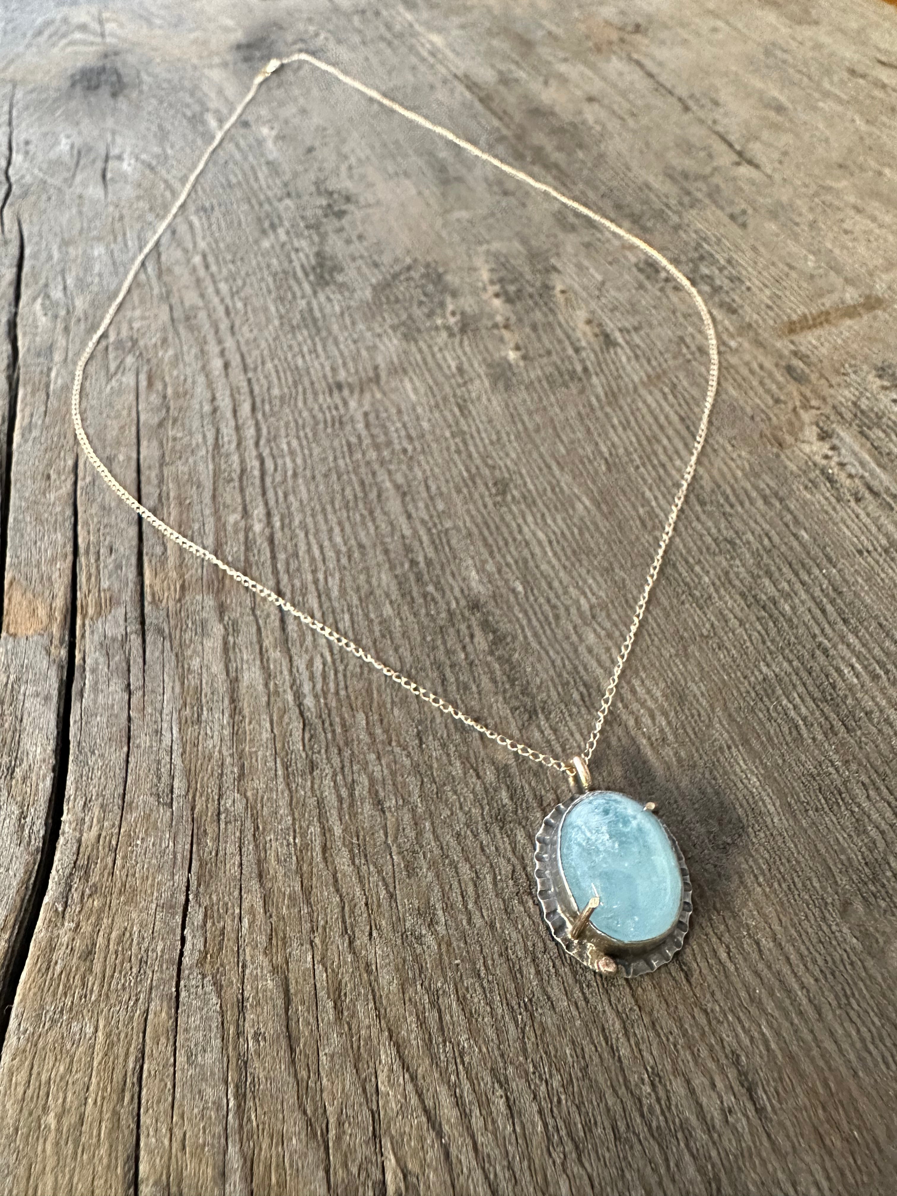 Blue Aquamarine oval stone set in silver with gold prongs and a small gold ball in the lower left corner on a gold chain laying on a wooden background.
